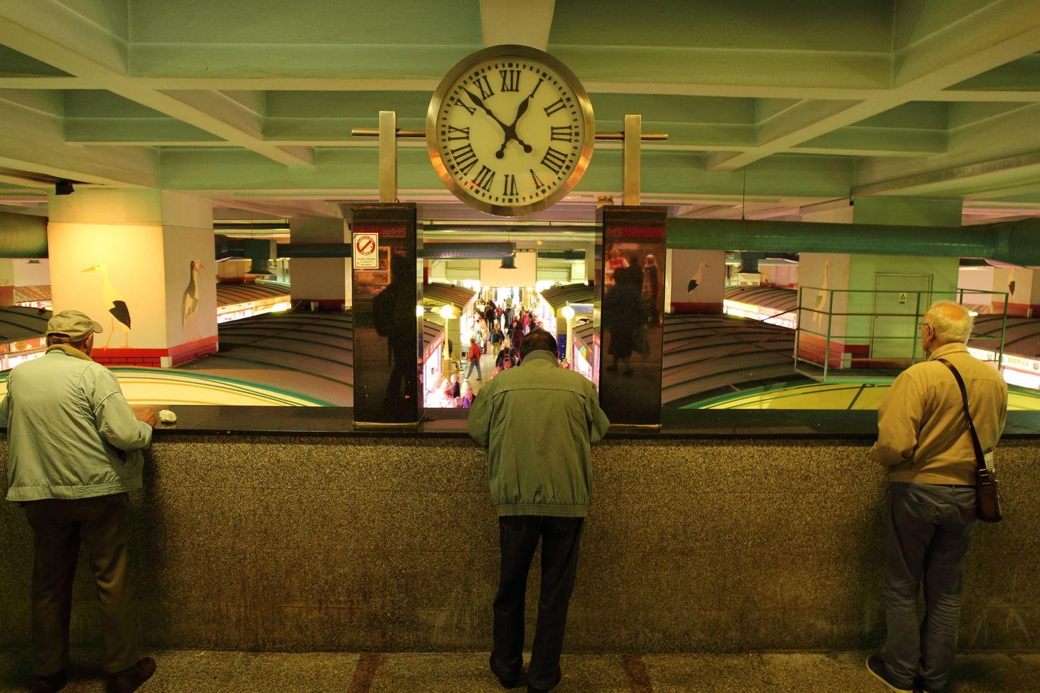 Interior of a subway station with a large clock and waiting passengers at the turnstiles.