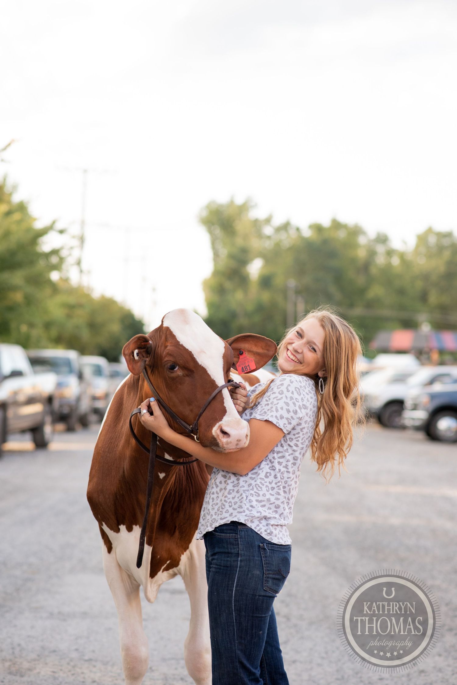 A brown and white cow gets hugs from someone in a floral top in a parking lot setting.
