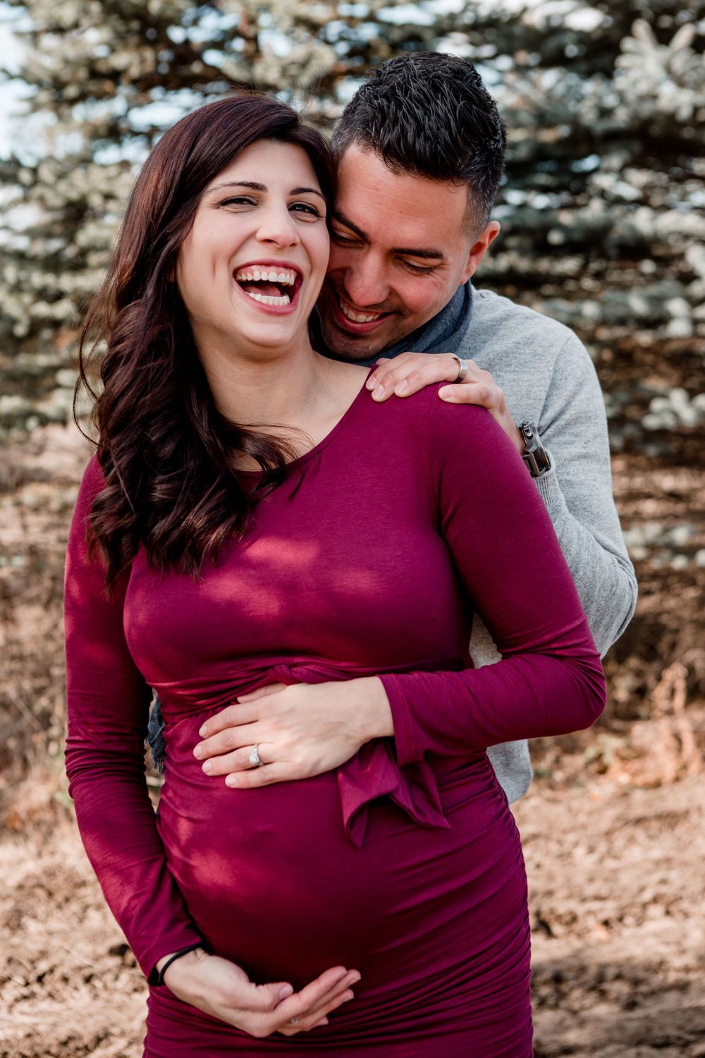 Joyful moments captured during a fall maternity session with couple embracing near pine trees.