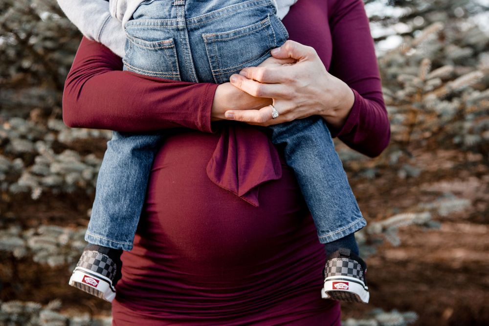 An intimate maternity photoshoot features loving hands cradling a baby bump in a burgundy dress against an autumn backdrop.