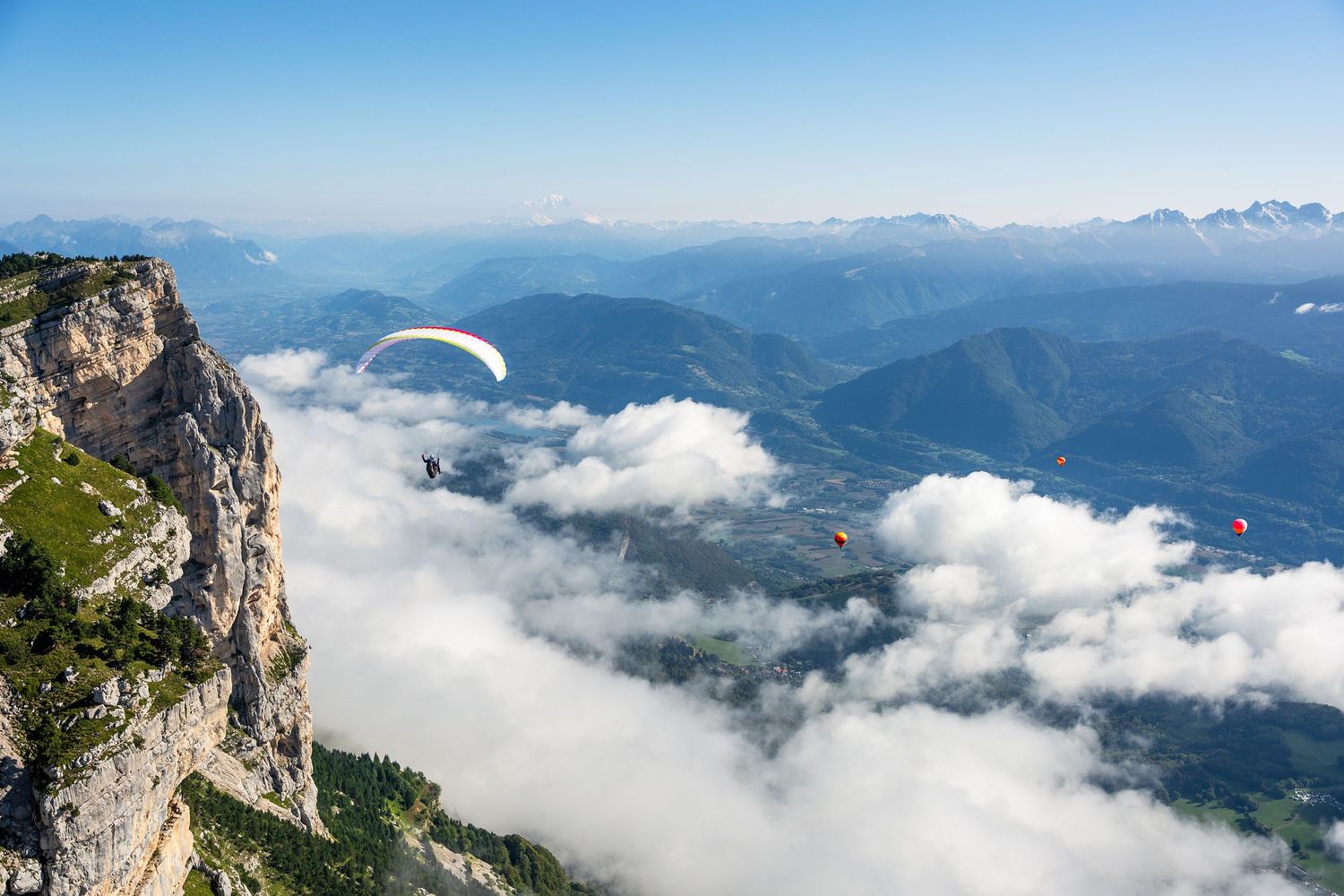 Two paragliders soaring above misty mountain cliffs and valleys with dramatic cloud cover.