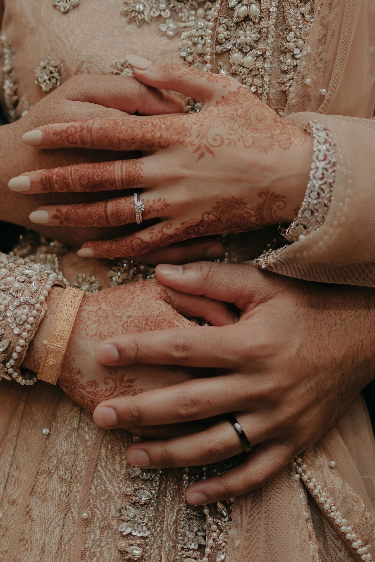 Close-up of hands with henna designs and wedding rings on embellished fabric.