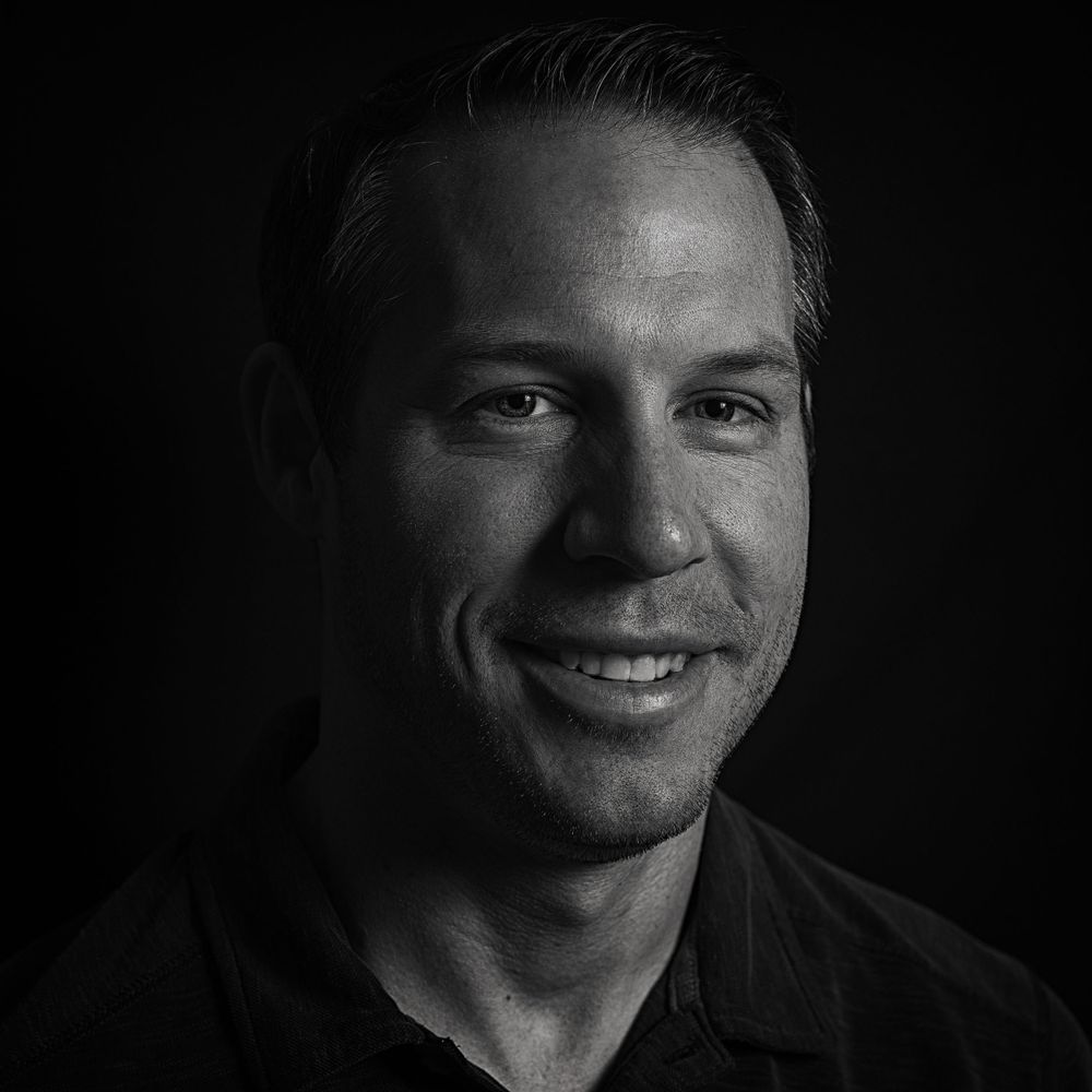 Black and white portrait of person in dark shirt smiling against black background.