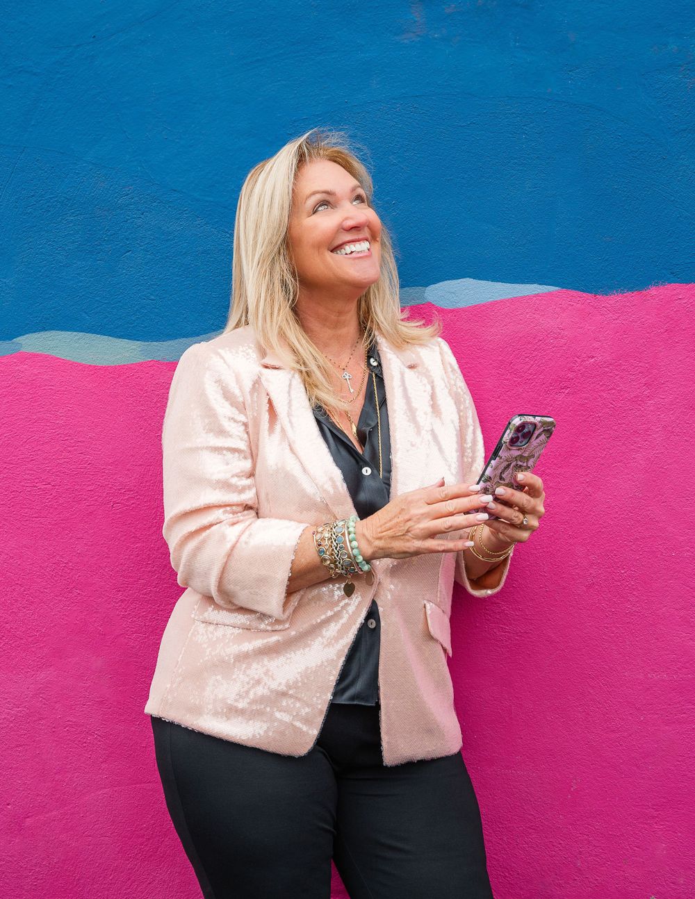 Woman in pink blazer using smartphone against bright pink and blue wall background.