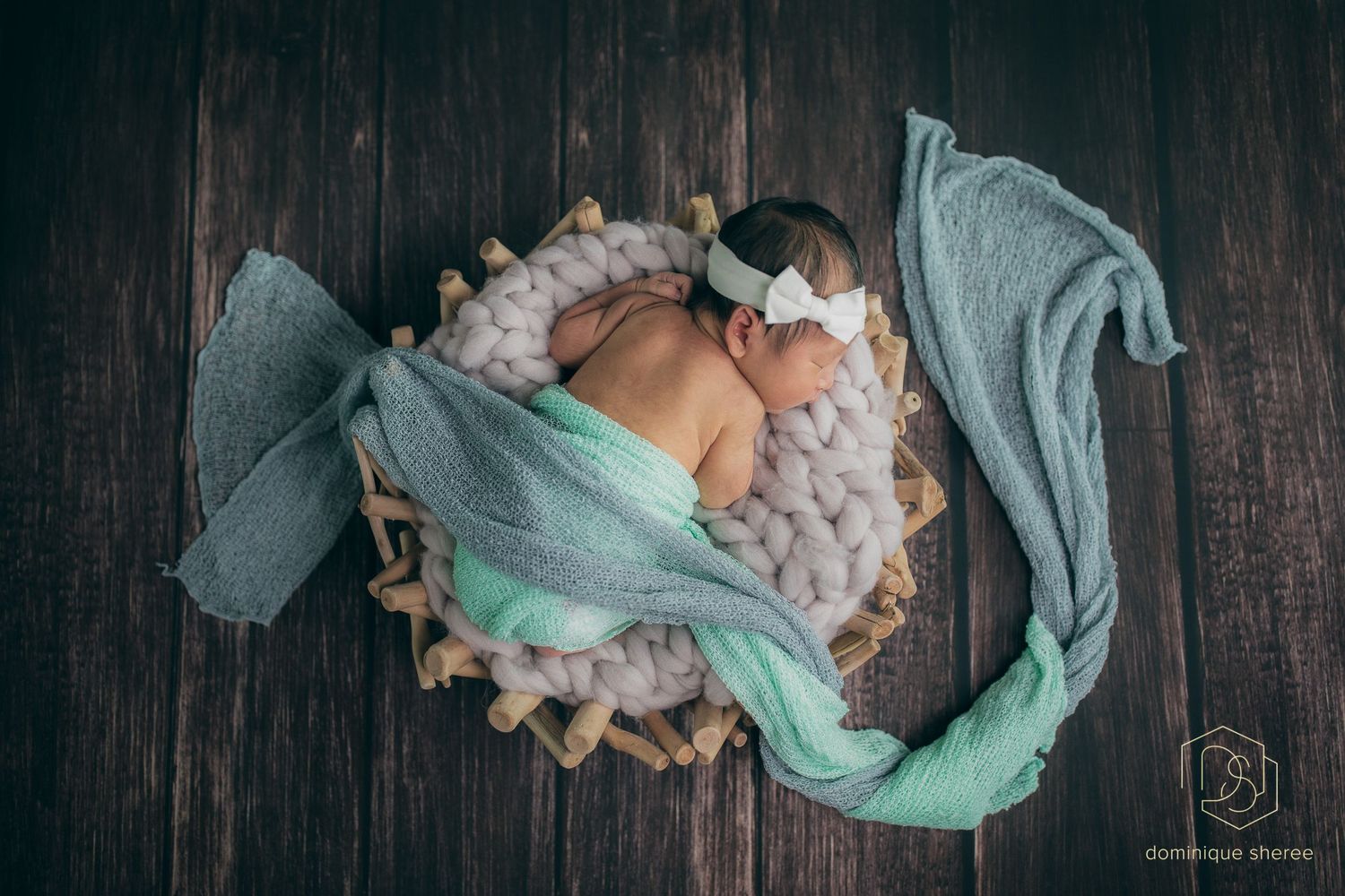 Newborn baby sleeps peacefully in mint green wrap on wooden backdrop with soft textures.