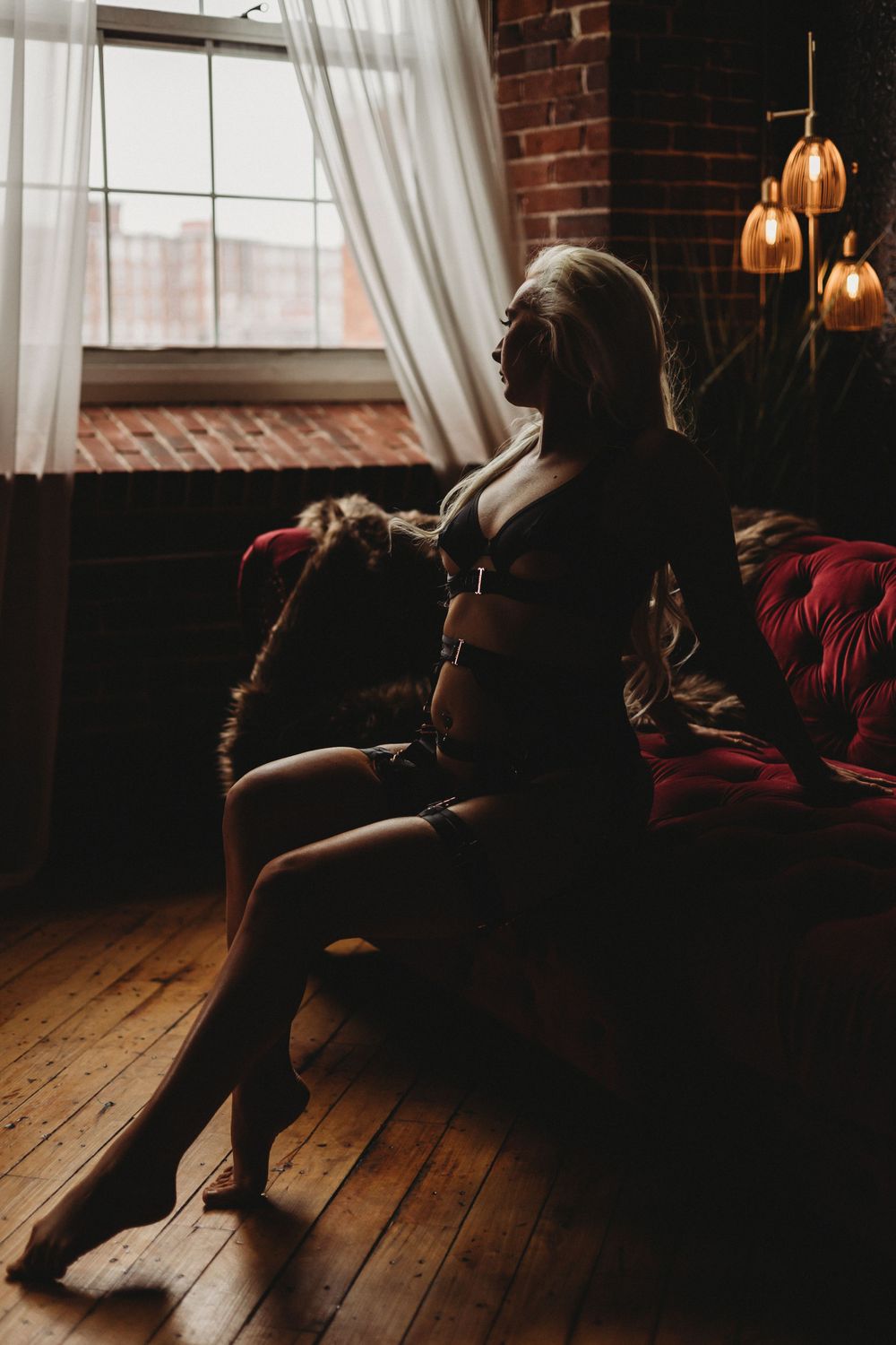Natural light boudoir portrait by window with industrial pendant lights and brick walls.