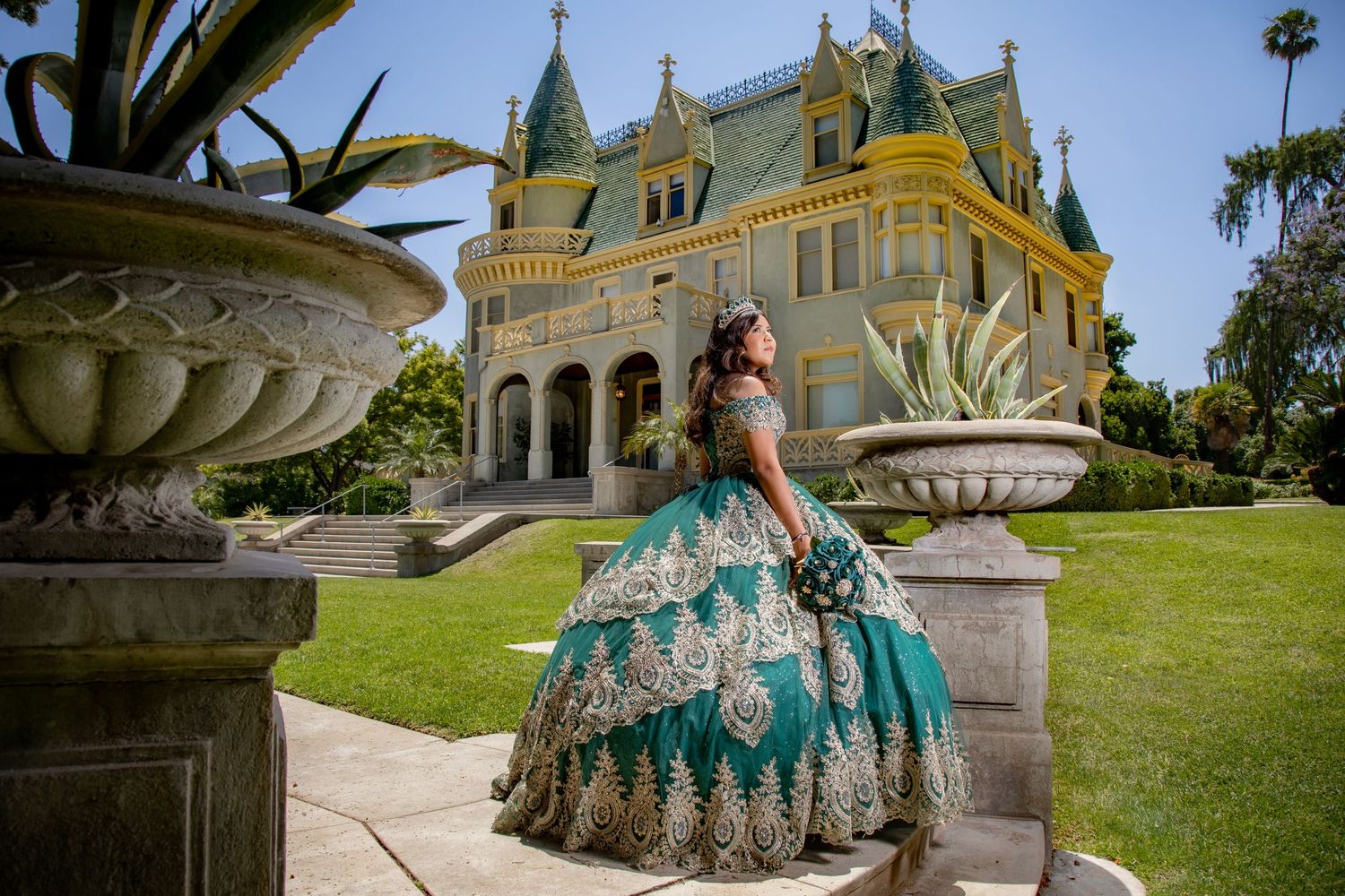Young woman in a teal and gold quinceañera gown poses in front of a Victorian mansion.