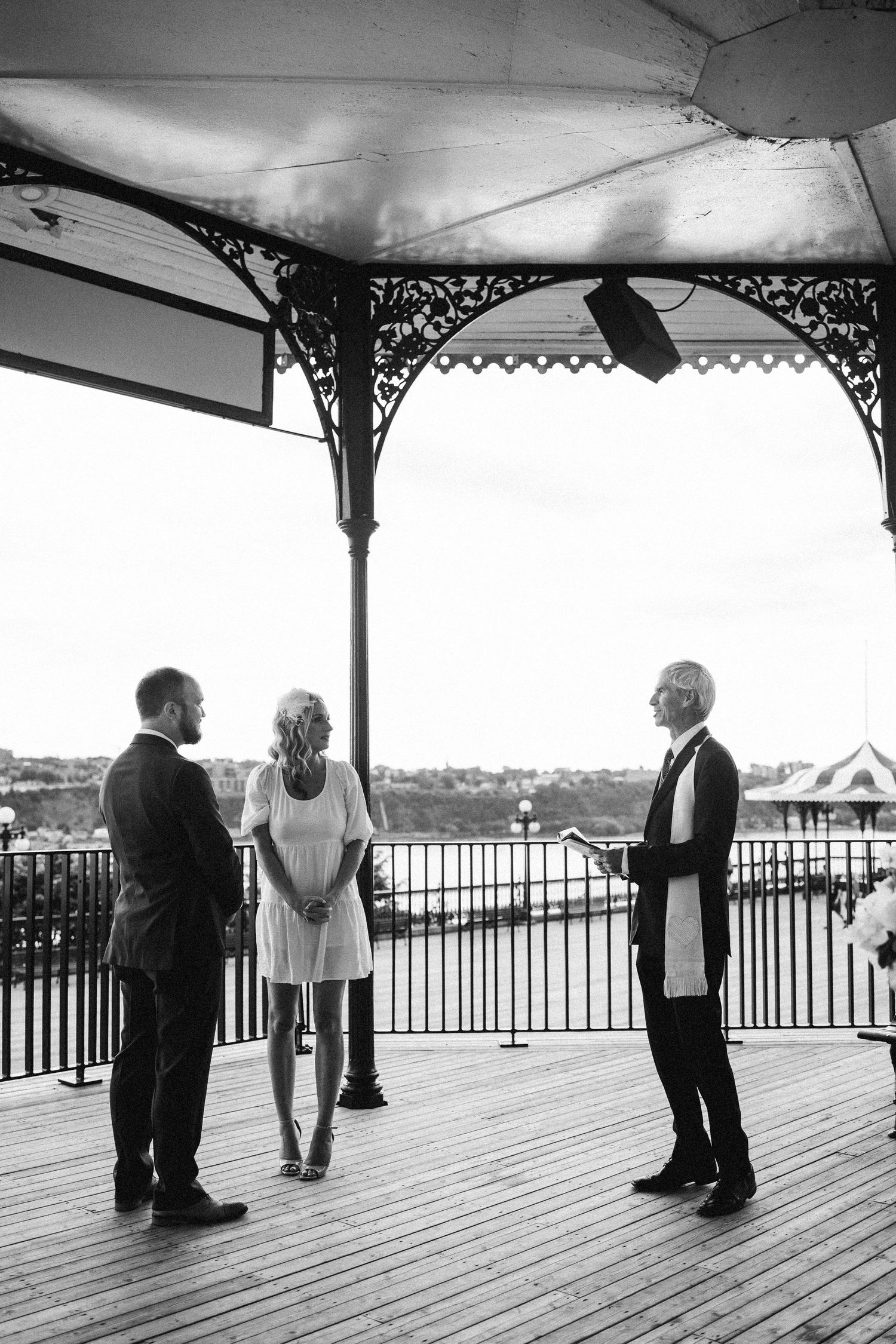 A black and white ceremony photo under an ornate gazebo overlooking water.
