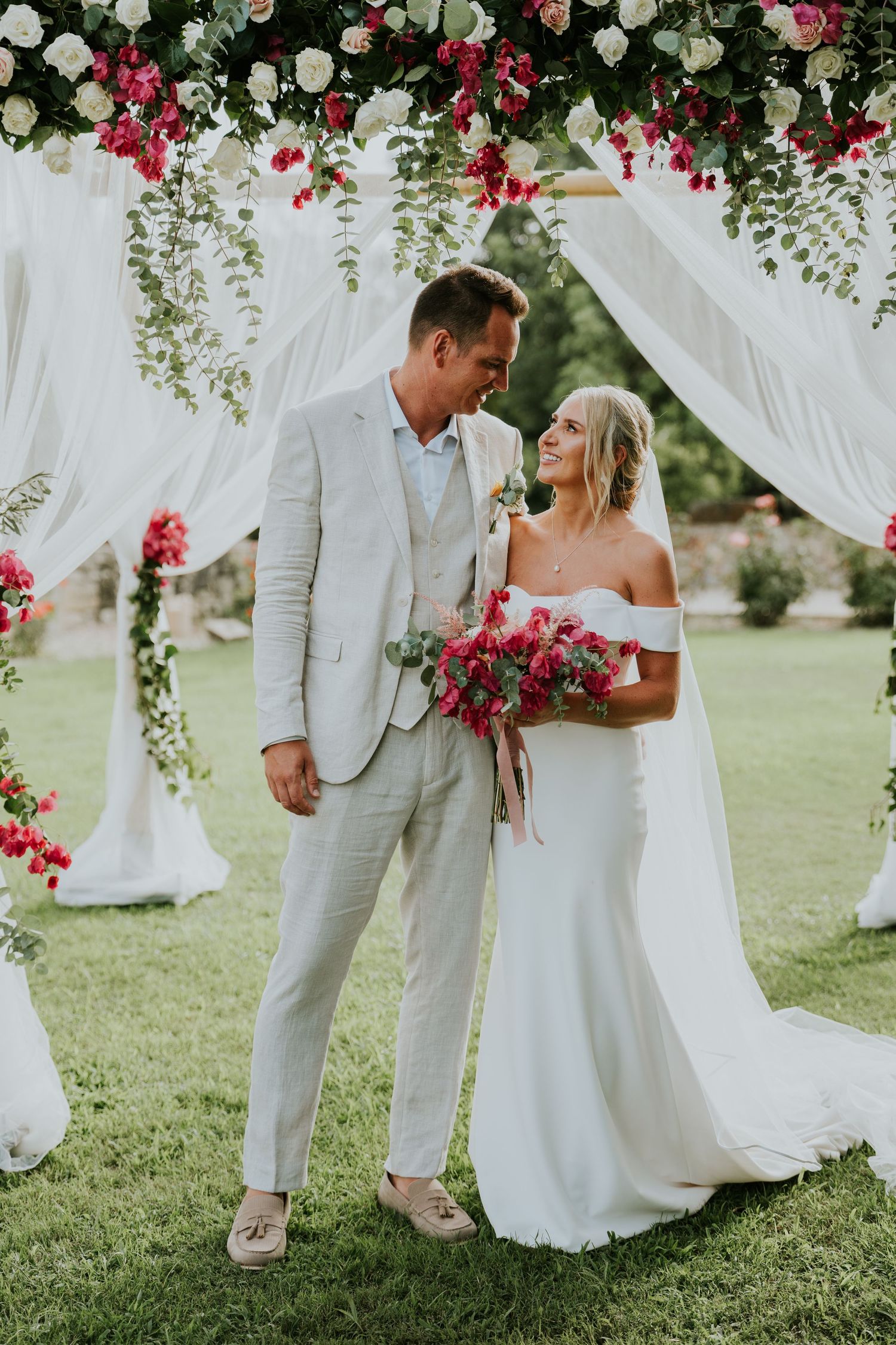 Wedding couple posing under floral arch with red and white flowers and flowing fabric.
