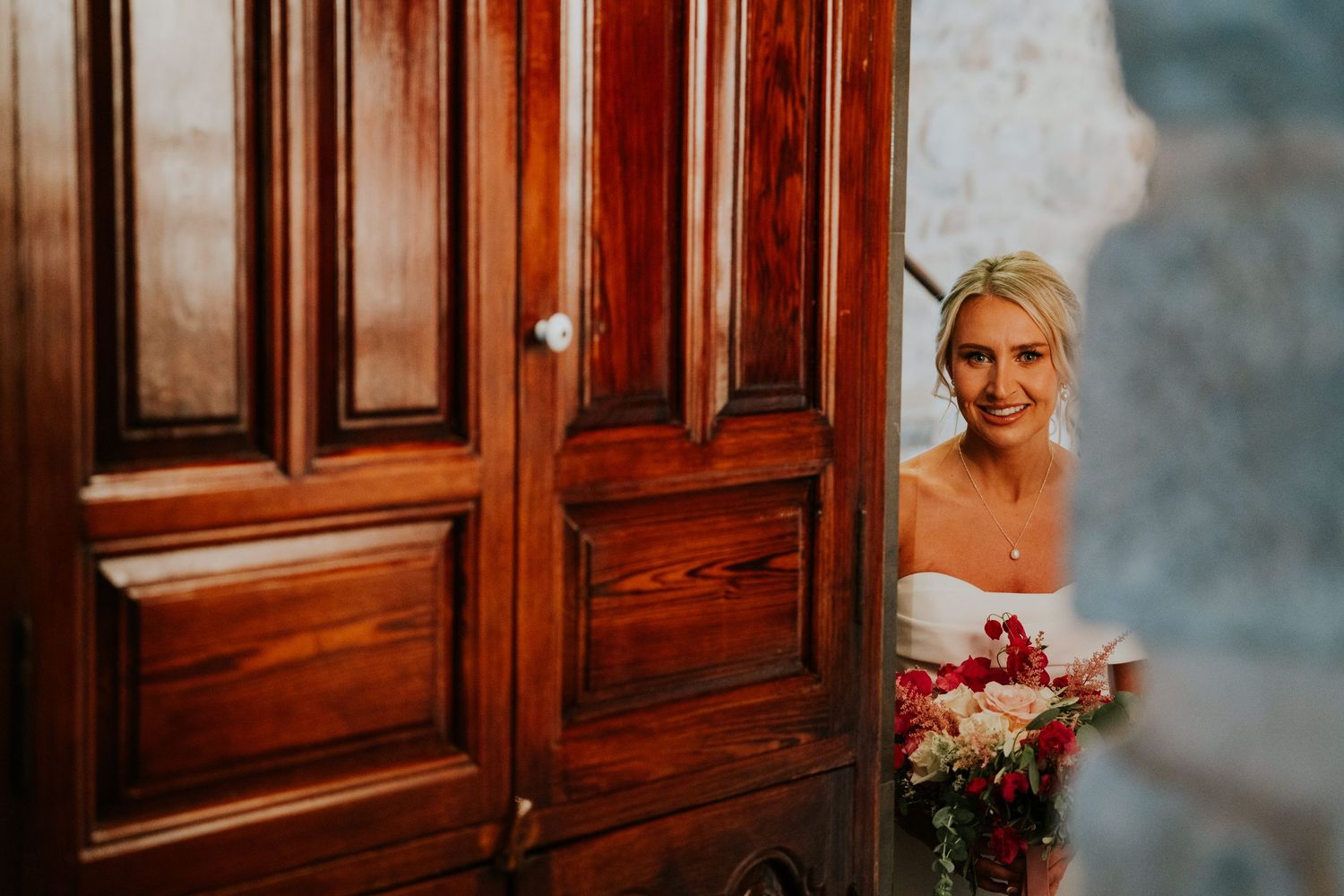 A peek through wooden doors reveals a bride holding a red and white bouquet.