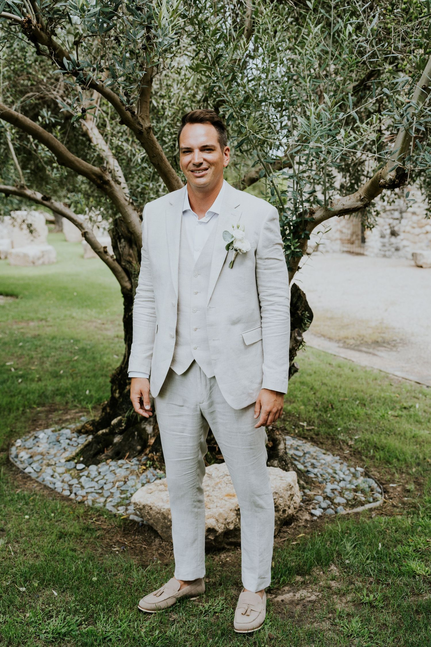 A groom in a light gray suit standing in front of an olive tree in a garden setting.