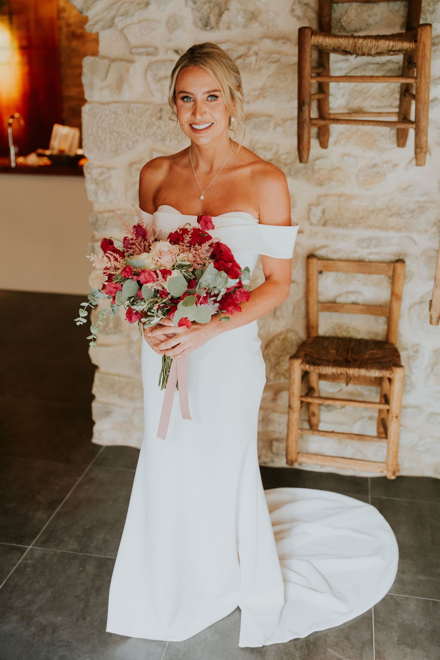 A bride in an elegant off-shoulder white dress holding a vibrant red and pink bouquet.