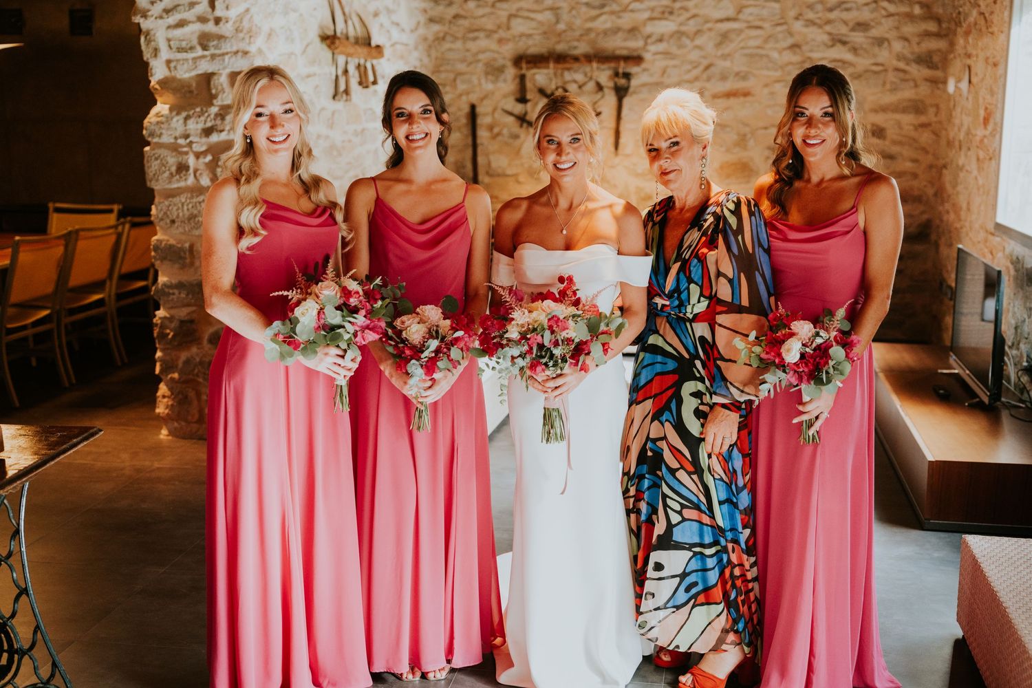 Bridal party in pink dresses posing indoors against a stone wall backdrop.
