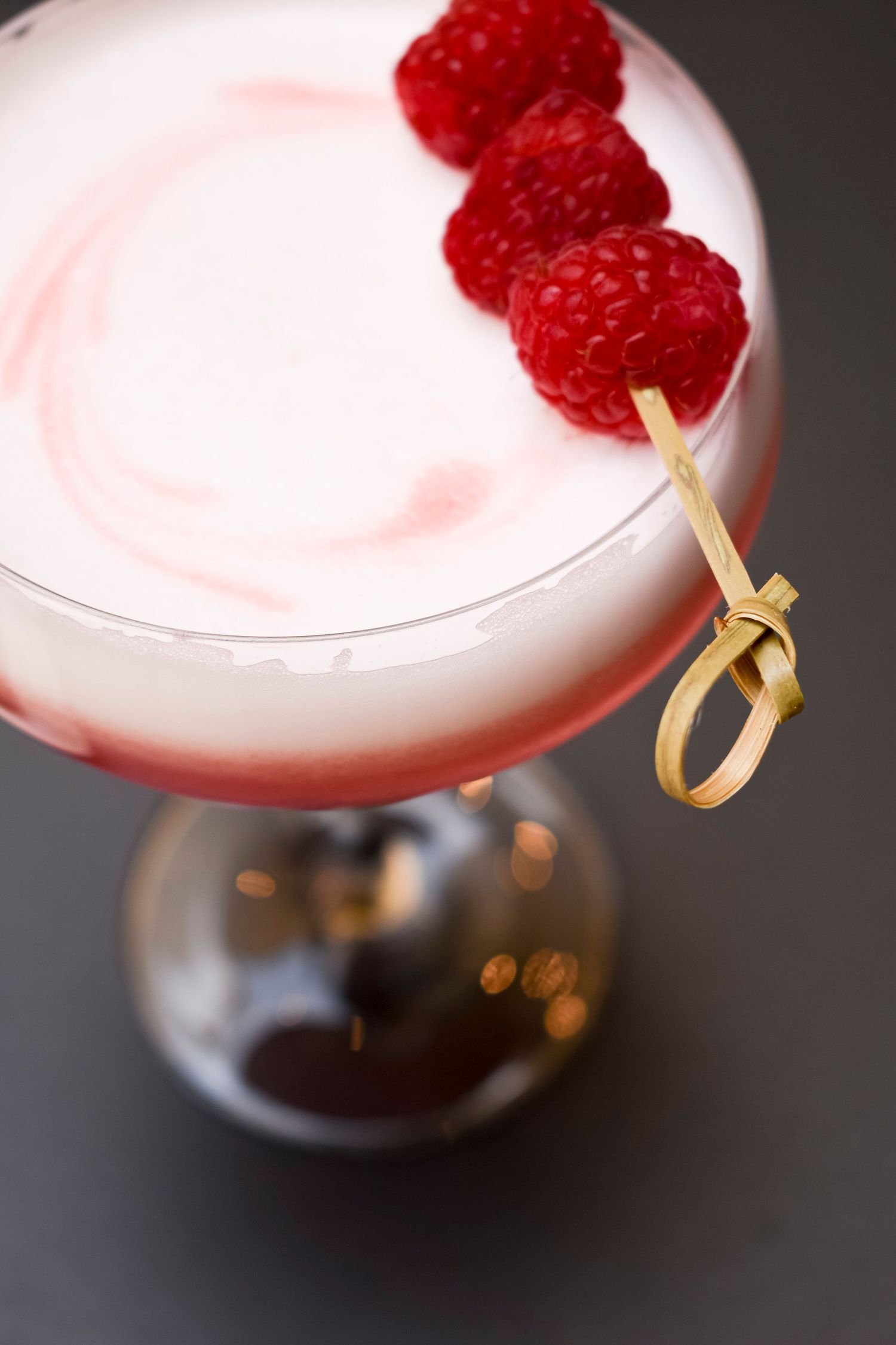 Pink cocktail garnished with fresh raspberries and golden bamboo pick.