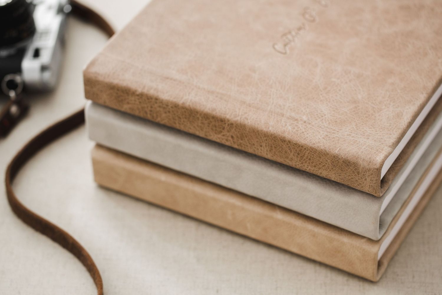 Stack of three vintage leather bound books with brown leather camera strap visible in corner against light background.