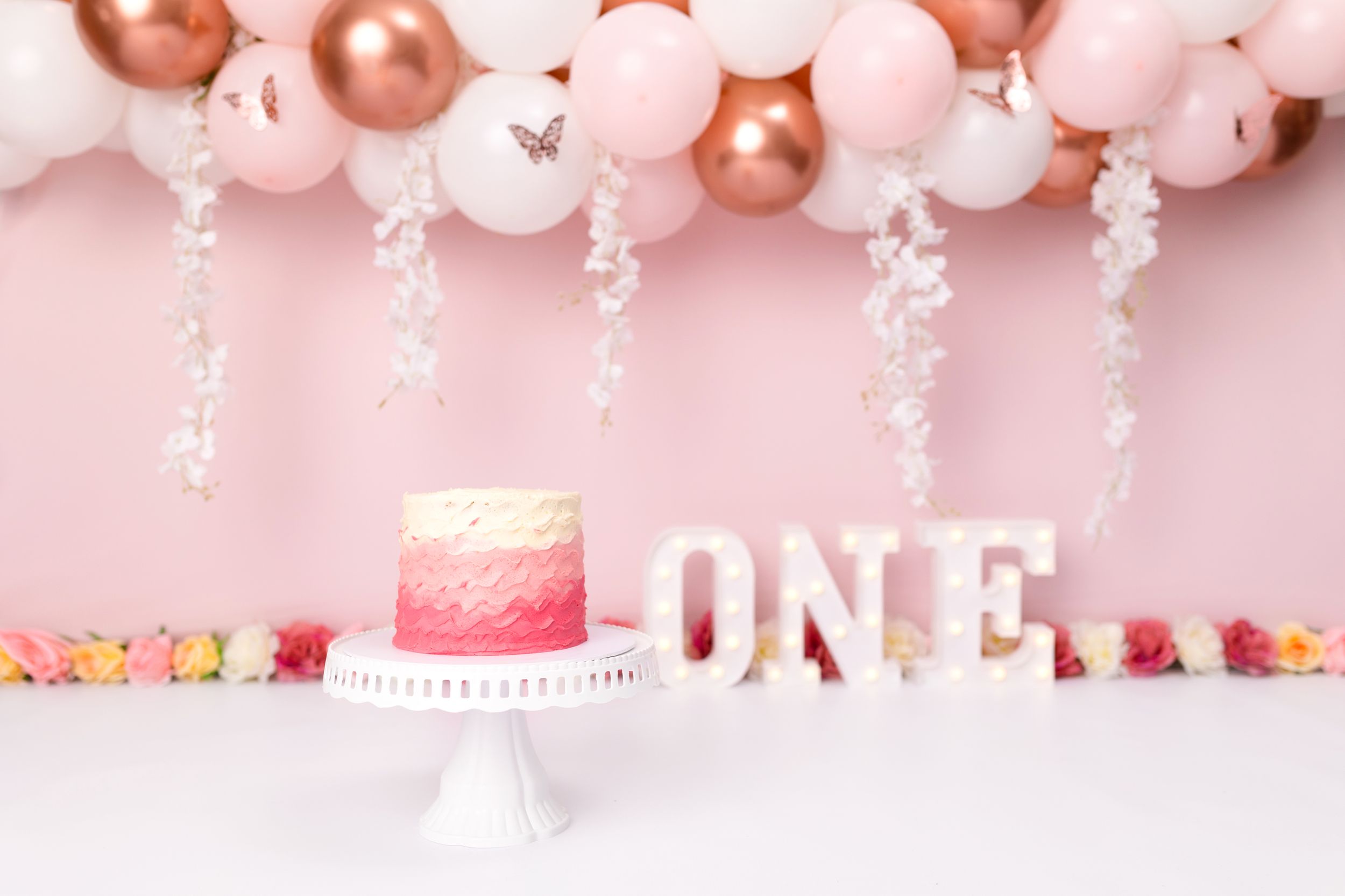 First birthday setup with pink cake and balloon garland against rose pink background.