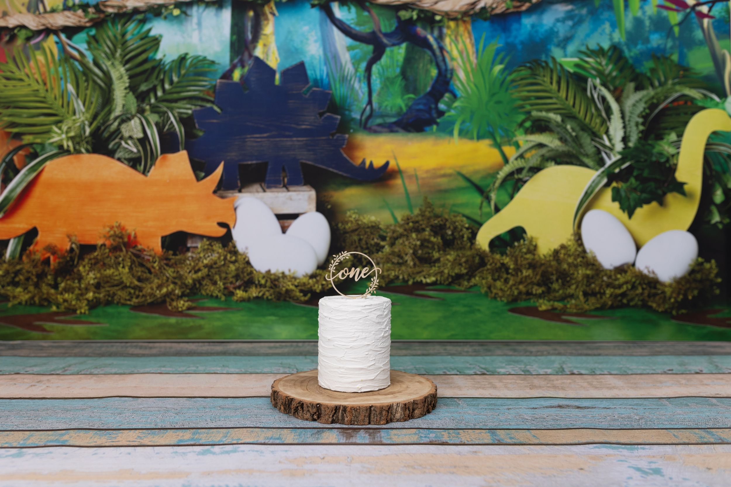 Decorative dinosaur-themed birthday cake setup with plush toys against a painted backdrop.