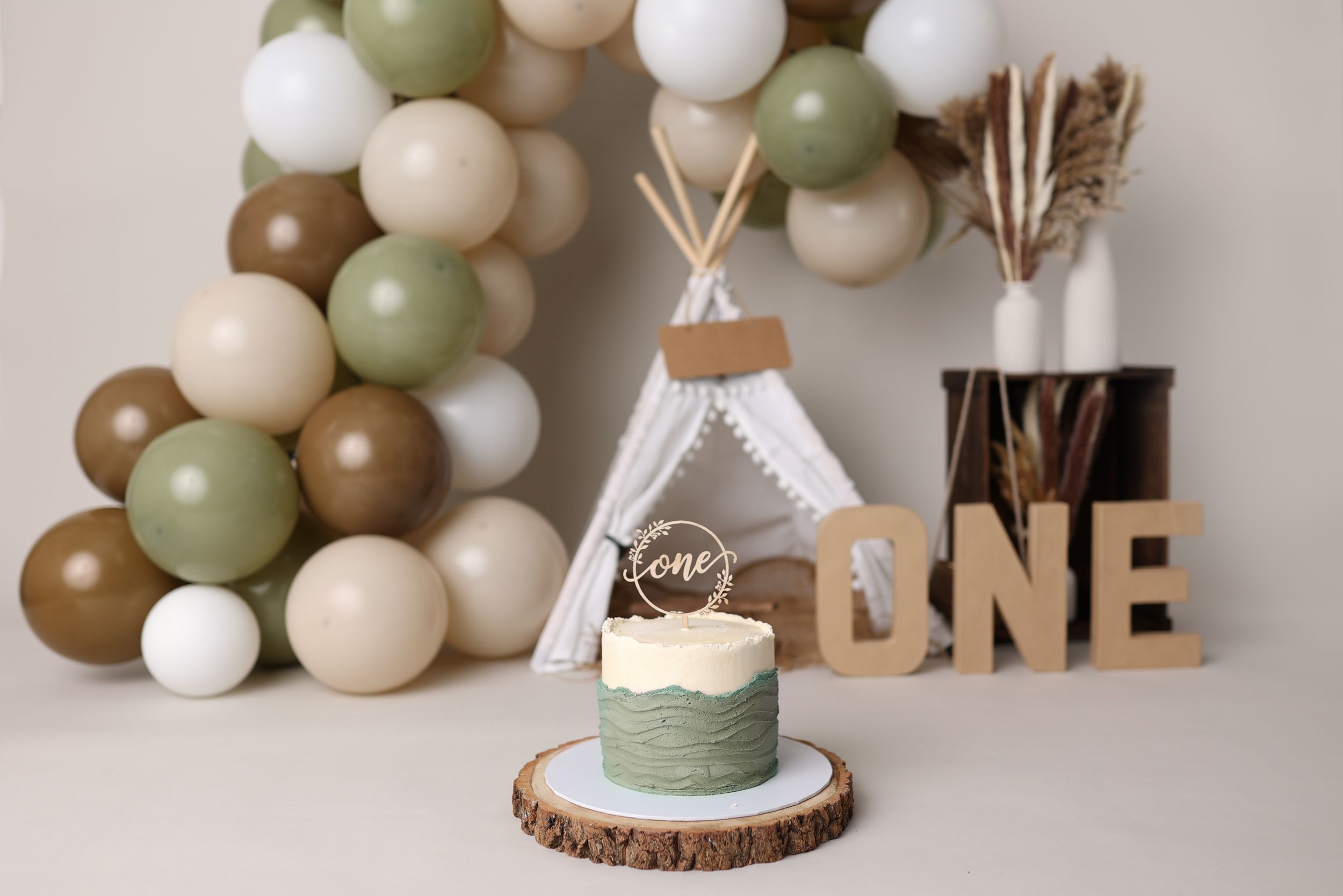 Rustic first birthday party decor with balloon arch, teepee and wood accents in neutral earth tones.