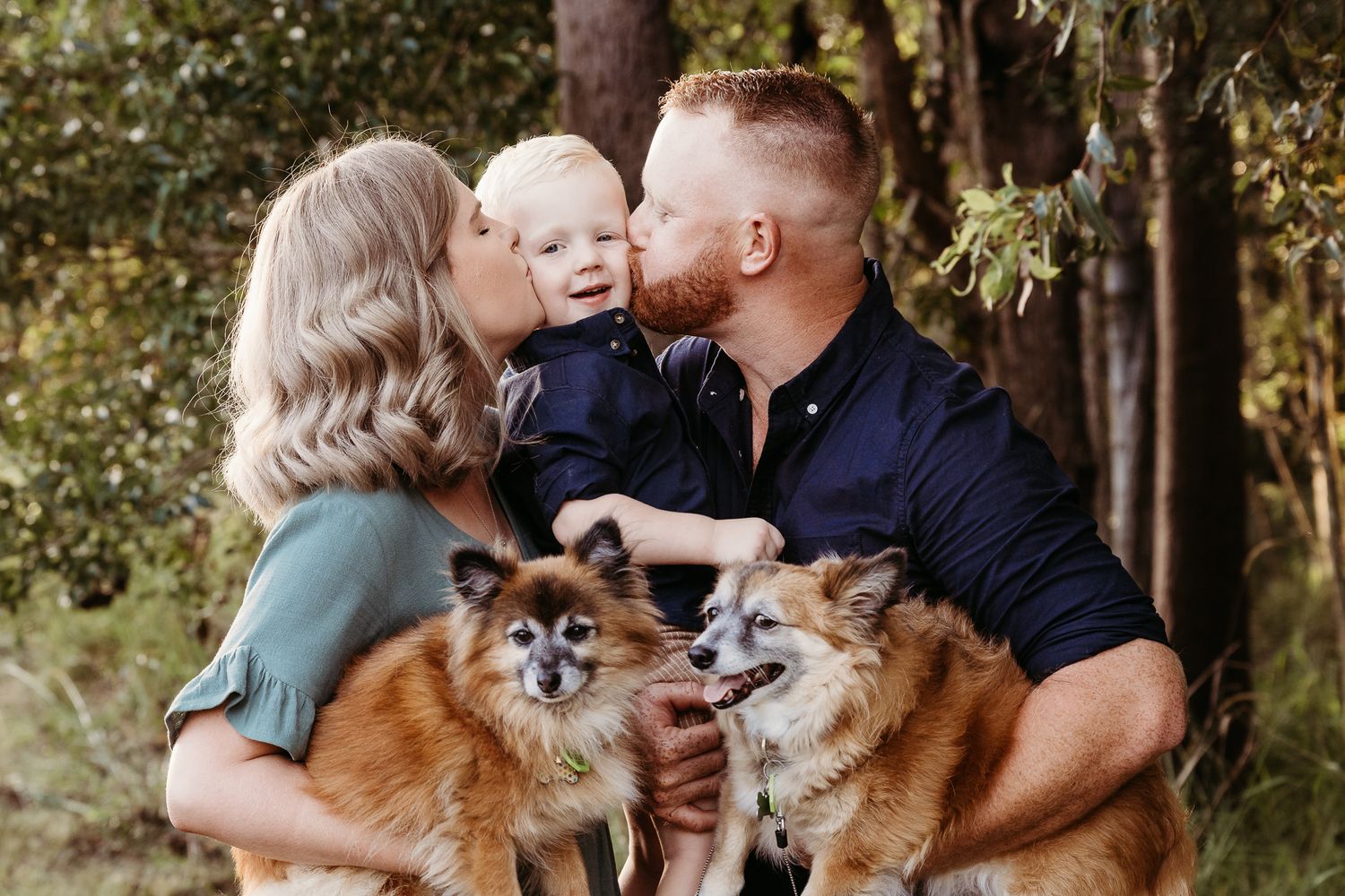 A family poses outdoors with their two fluffy Pomeranian dogs among trees.