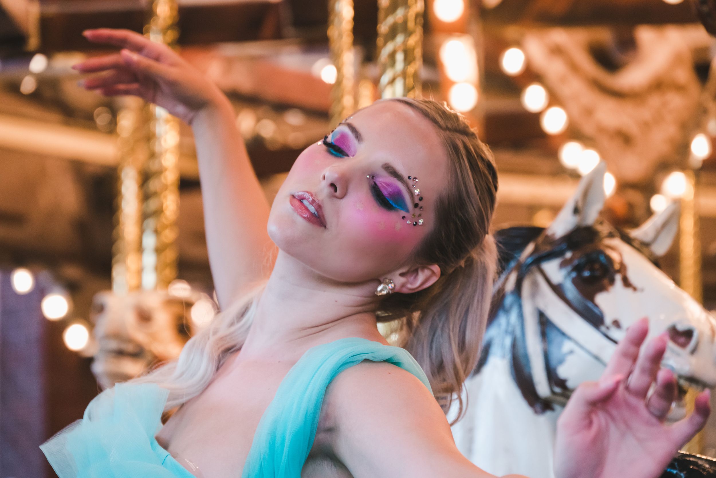 Young woman with colorful eye makeup and gems poses gracefully at a carousel.