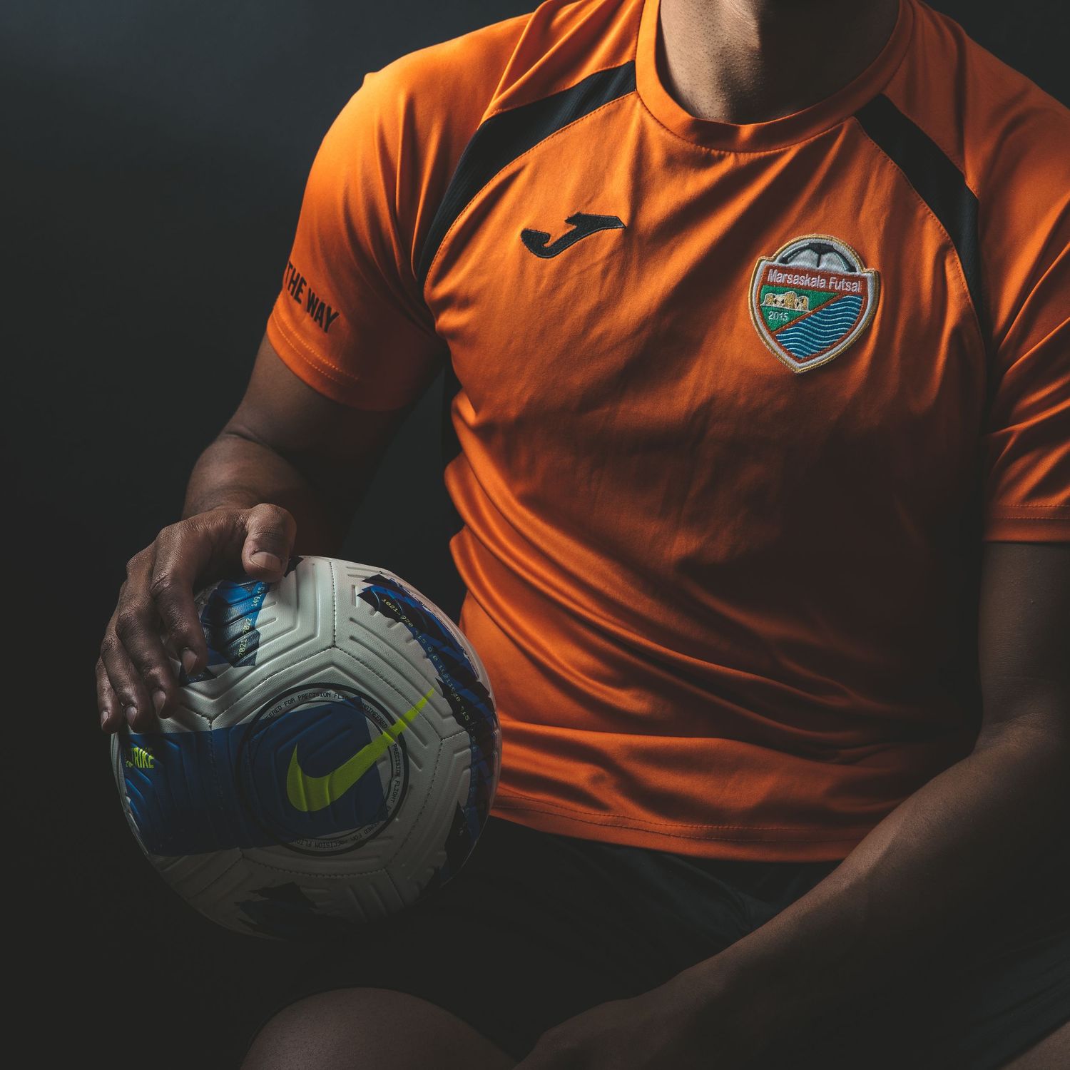 Soccer player in orange Joma jersey holding Nike football against dark studio background.