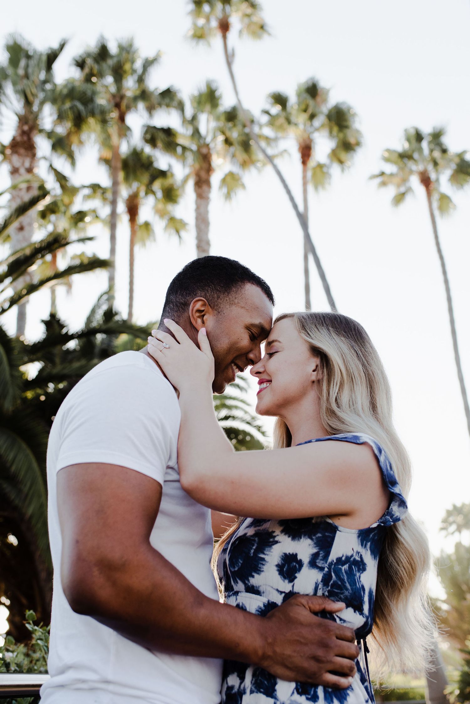 Couple stands with their foreheads together surrounded by palm trees and sun shining through behind them.
