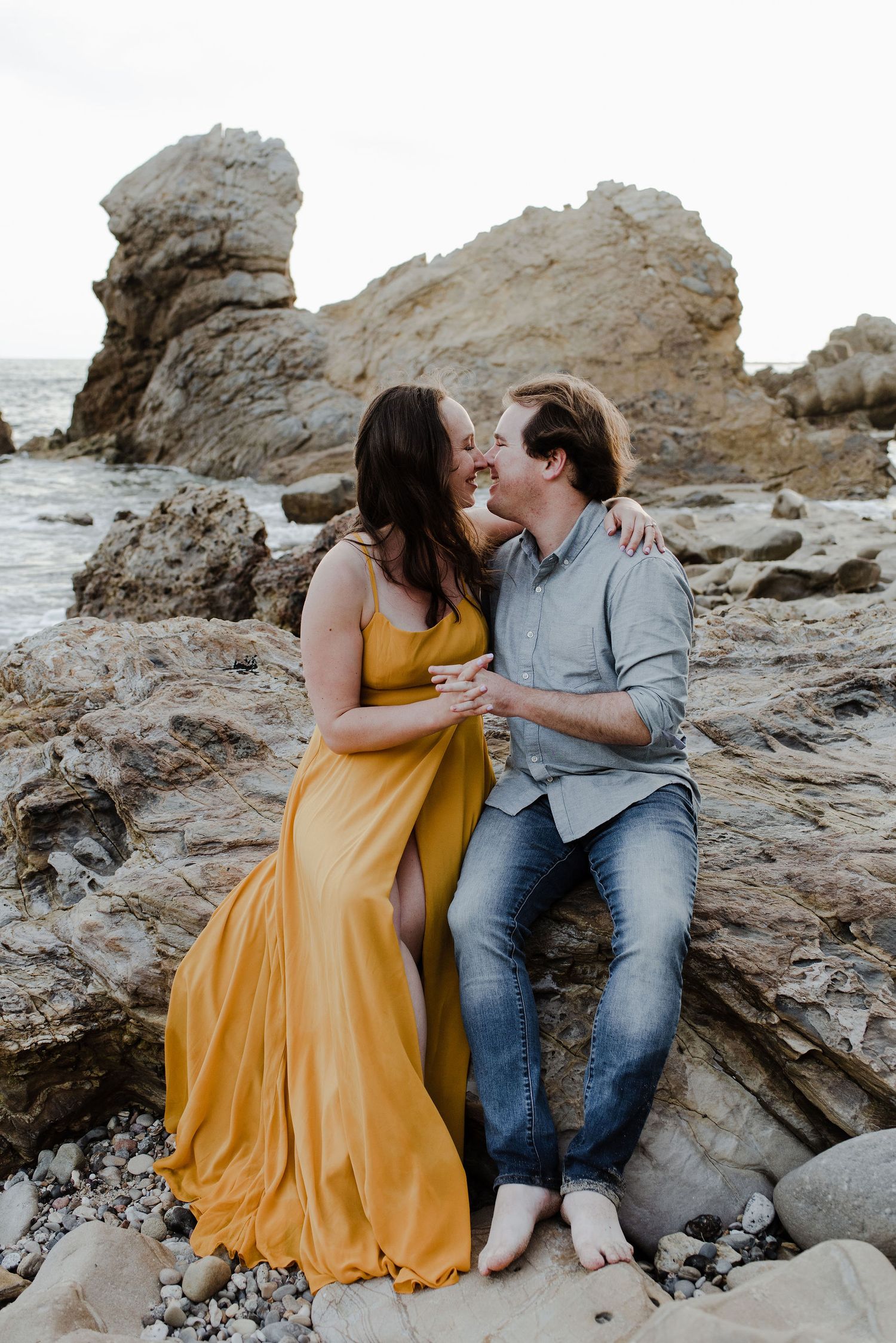 A laughing couple's noses are touching while sitting together and holding hands on a rocky beach in Southern California.