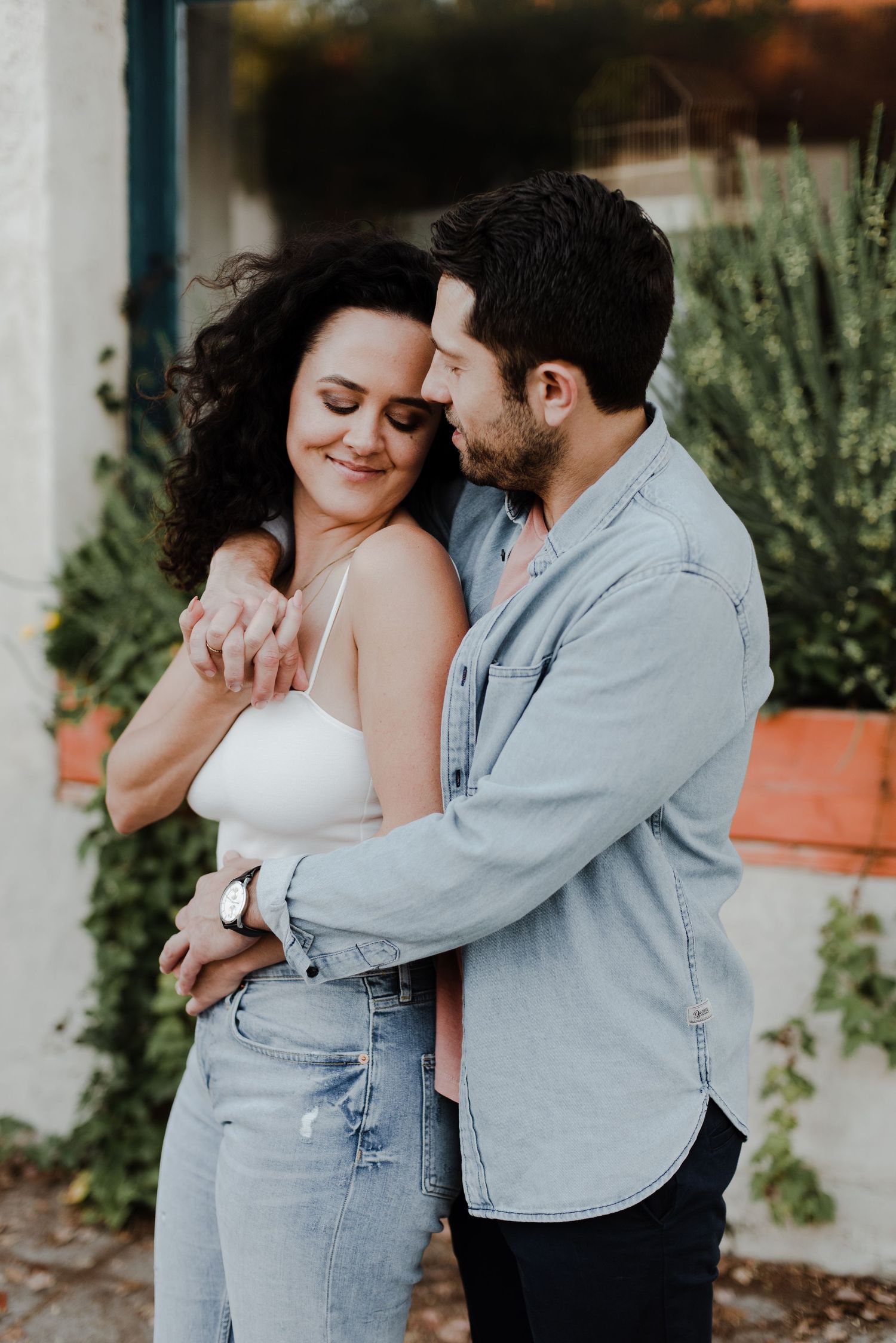 Man stands behind his fiancé, arms around her waist and shoulder as they lean into each other outside an antique shop.