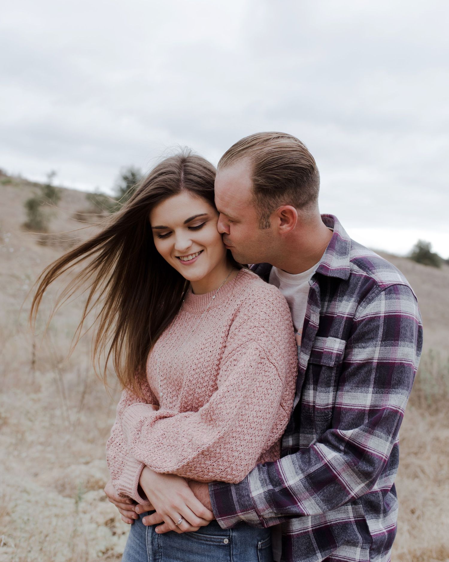 Man stands behind his fiancé, arms around her waist, leaning in to kiss her cheek as wind gently blows her hair