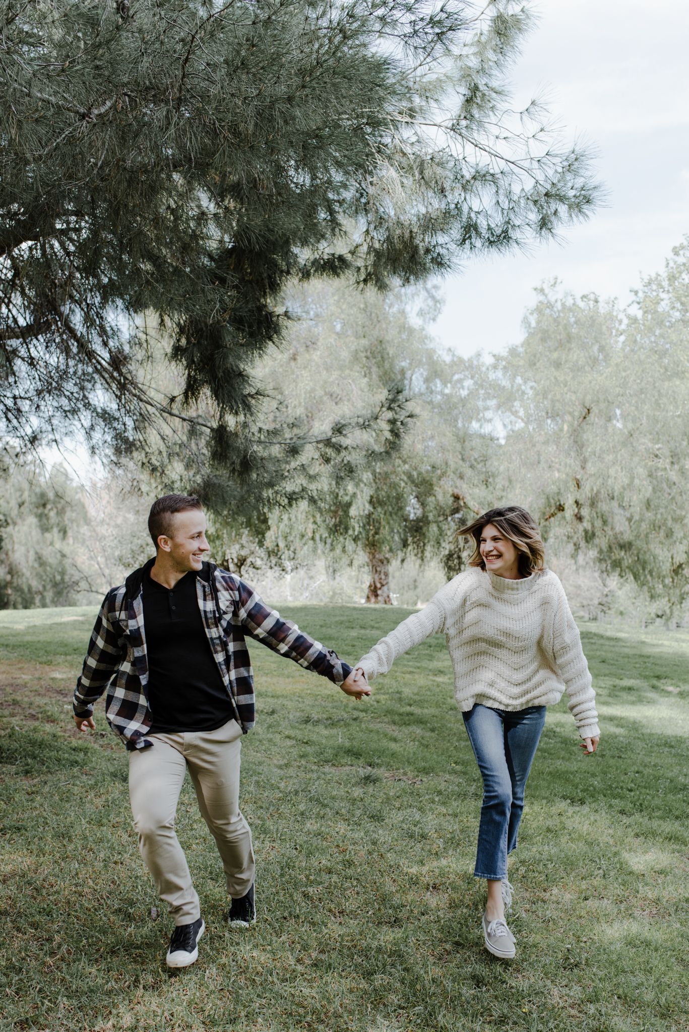 Couple holds hands looking at each other as they run toward toward the camera in a grassy park full of trees.