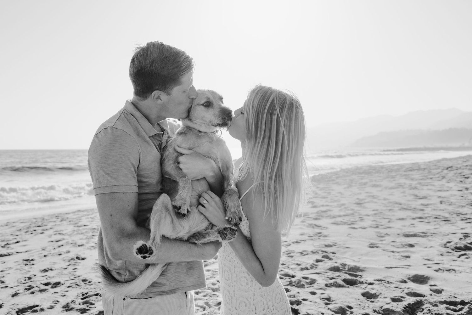 A couple on a beach holds their dog between them, both kiss the dog while the dog licks the woman's face.