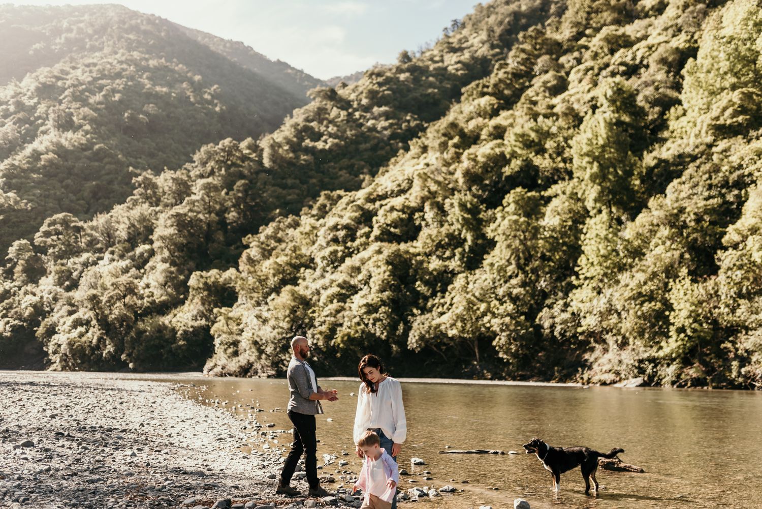 Family walking along a riverbank with mountains and a dog in the background.