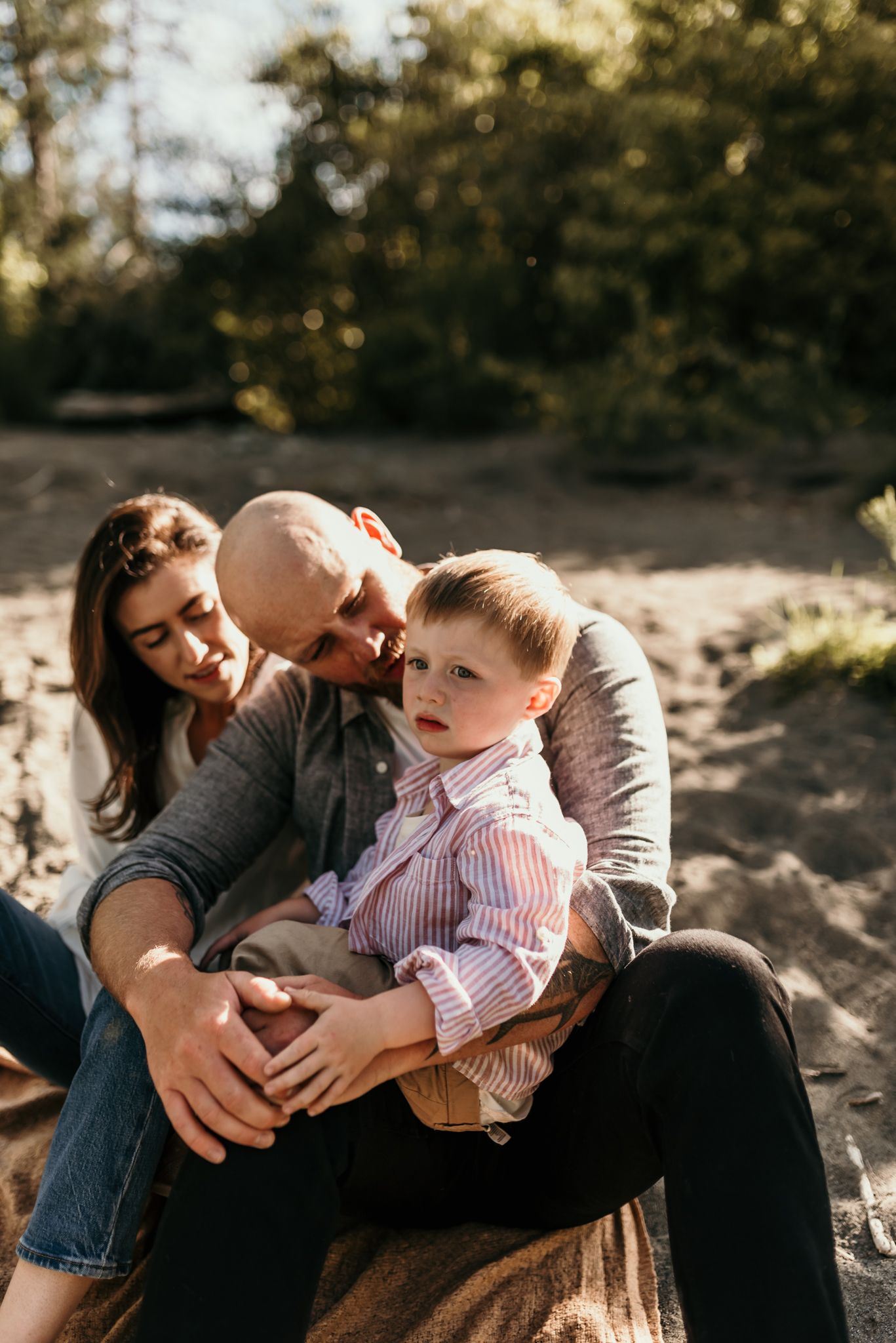 Family cuddles together on blanket in warm sunset lighting among trees and greenery.