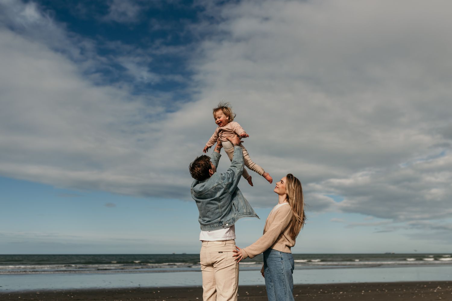 Joyful family moment at the beach with lifted child against cloudy sky and ocean backdrop.