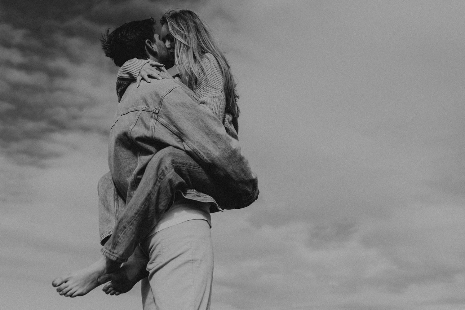 Romantic black and white portrait of a couple embracing against cloudy sky.