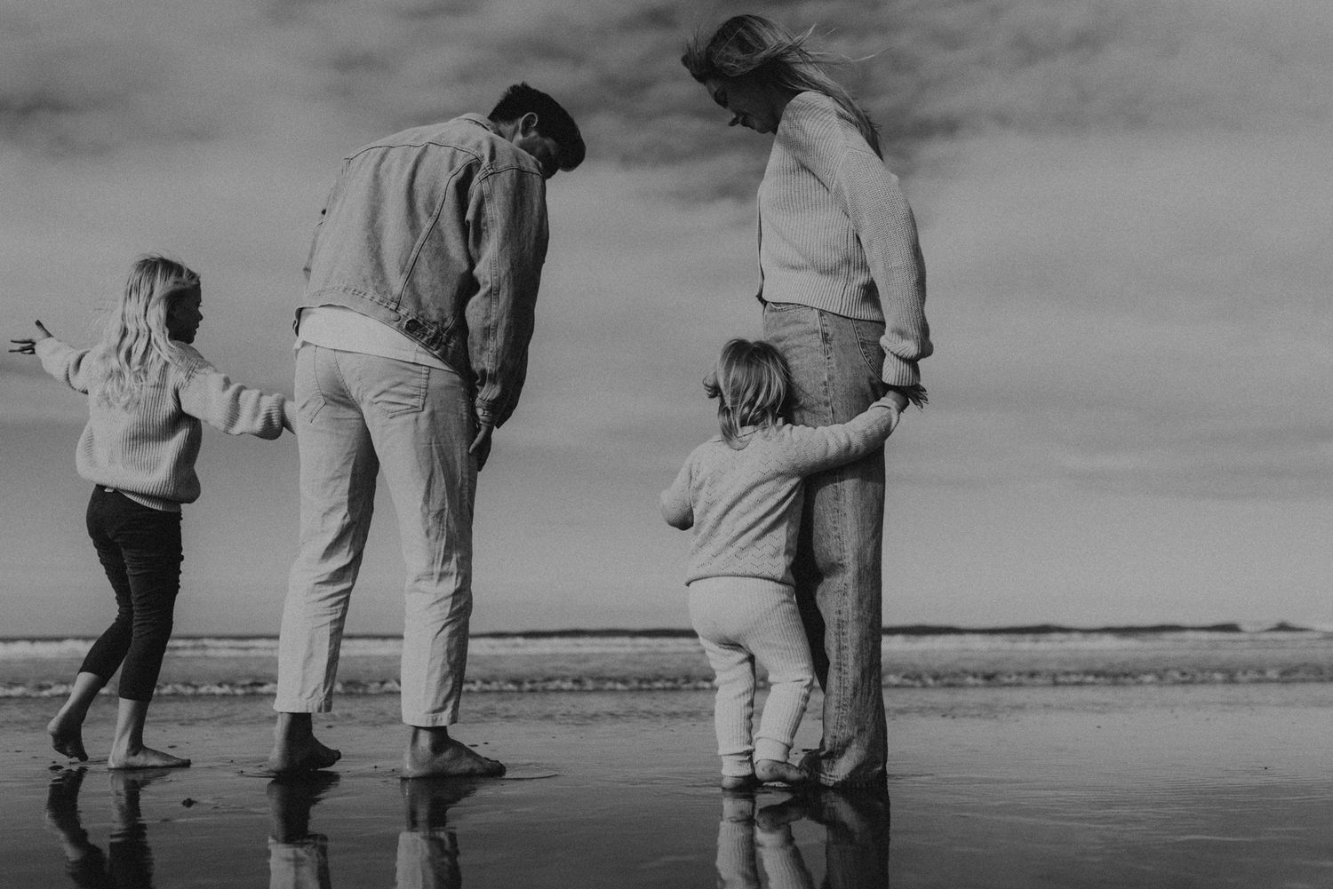 Family exploring and playing together on a reflective wet beach during an overcast day.
