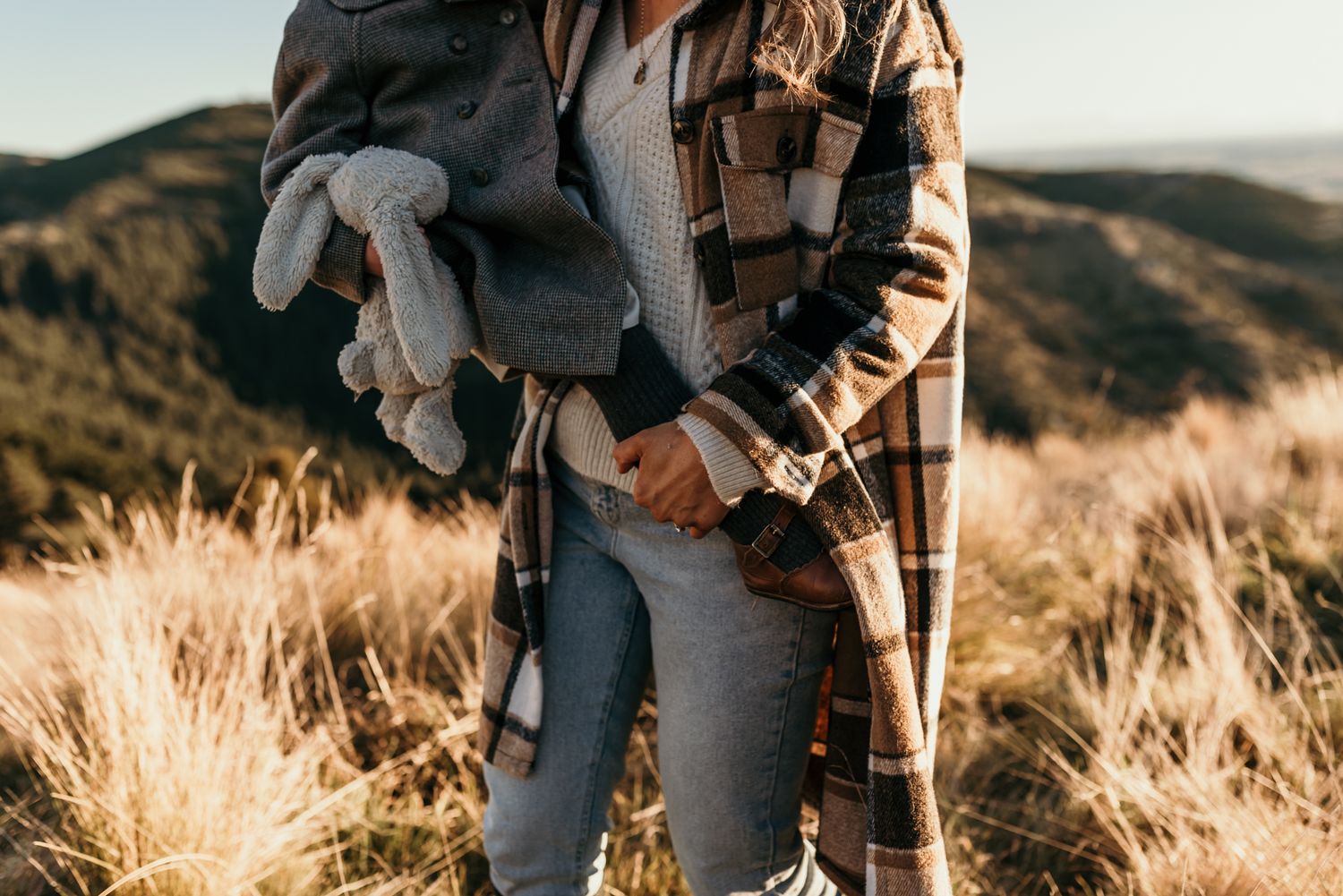 Person holding child while standing in golden grass on mountainside during sunset.