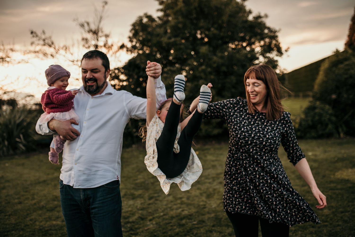Family playing together outdoors with a child being swung upside down during sunset.
