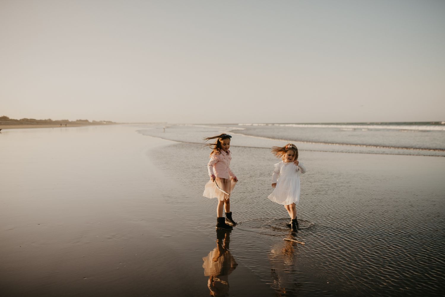 Two figures in white dresses playing at the water's edge during sunset.