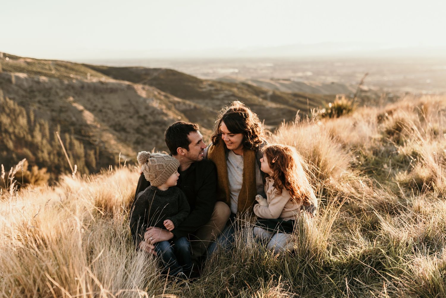 A couple sitting in golden grass on a hillside at sunset enjoy a tender moment together in nature.