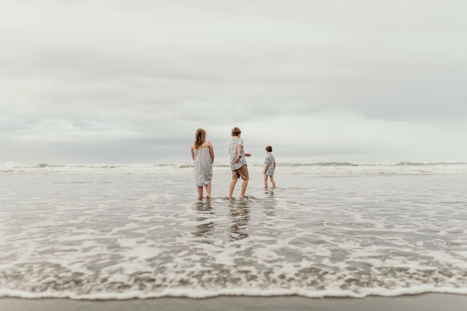 Three silhouettes walking through shallow ocean waves on an overcast beach.