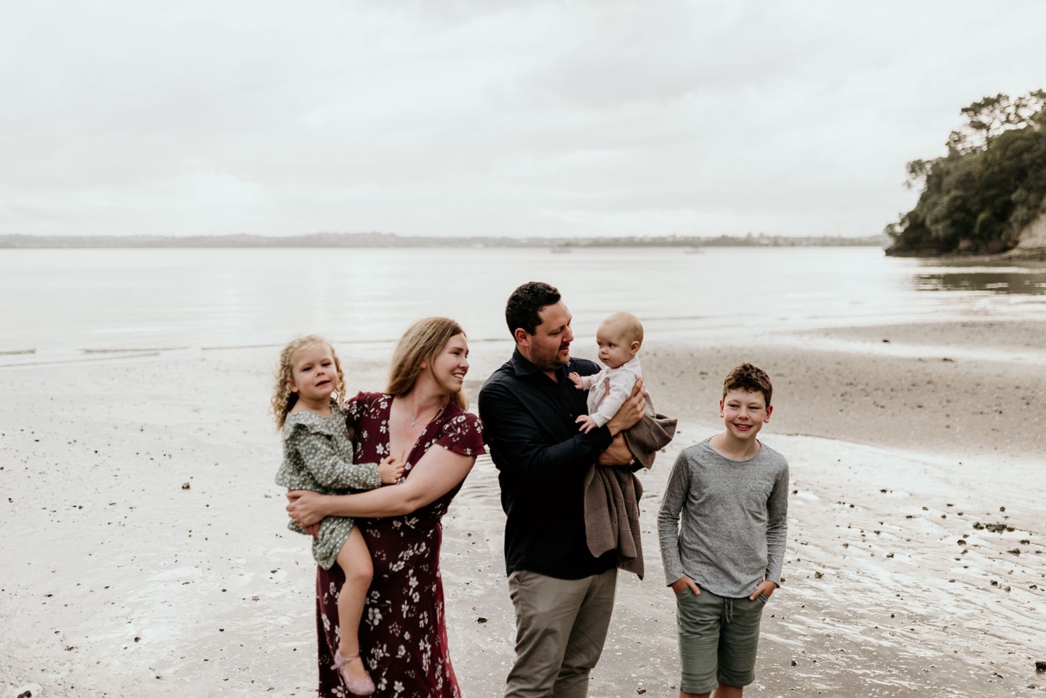 Family of five poses together on overcast beach with trees in background.