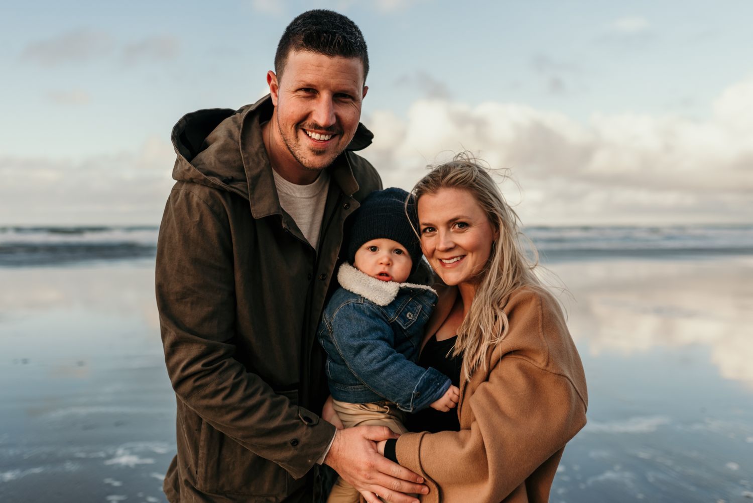 Winter beach portrait of family in warm coats with ocean waves and reflective sand in background.