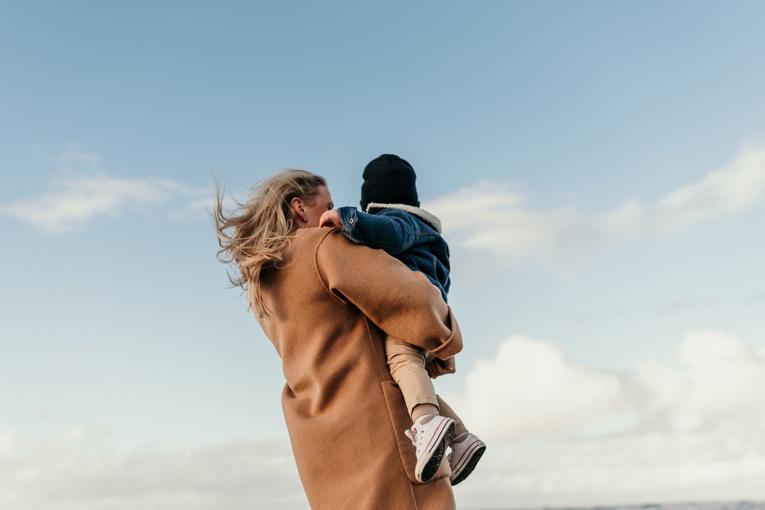 Parent lifting child against blue sky in camel coat.