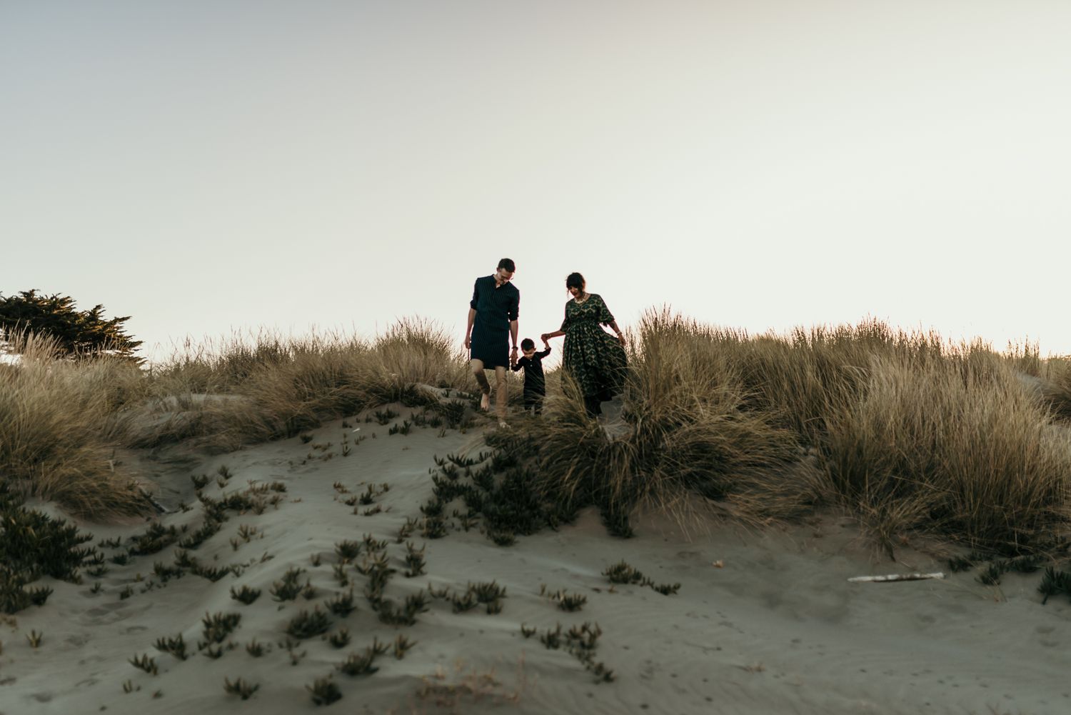 Family members climbing sand dunes together at dusk with wild grasses in silhouette.