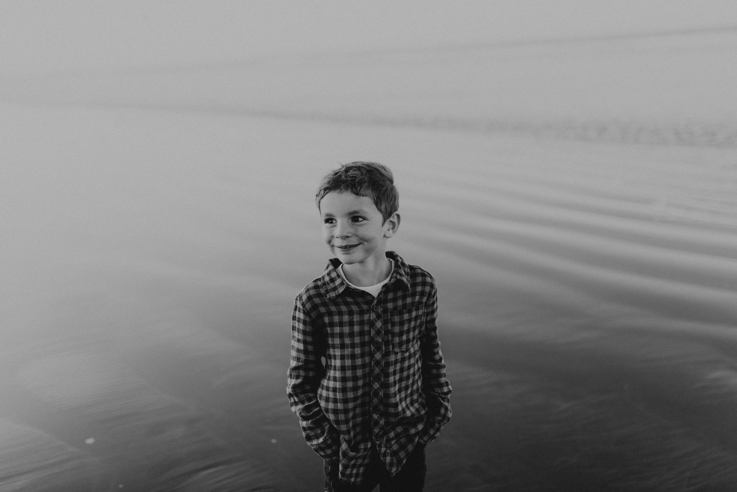 Black and white portrait of child in plaid shirt against misty water backdrop.