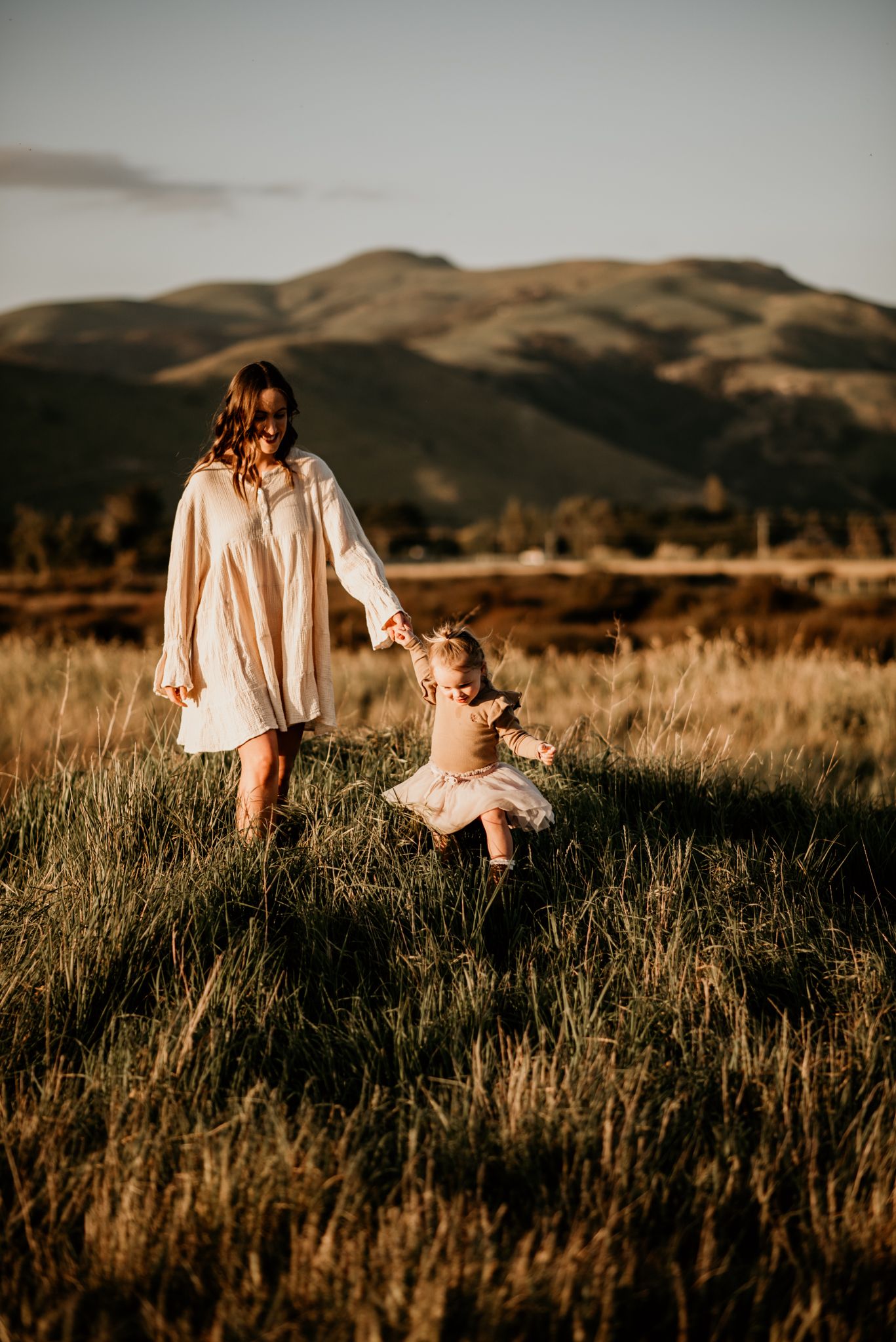 Two figures in white dresses dance together in a golden field at sunset with mountains in the background.