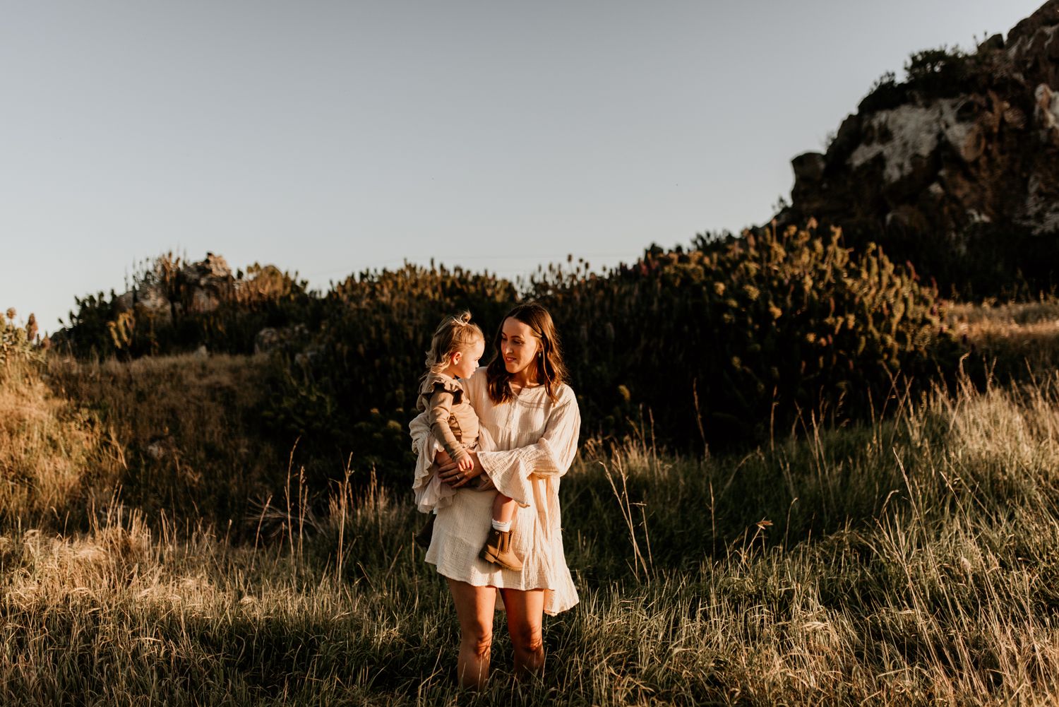 Three people share an intimate embrace while wearing white dresses in a golden meadow at sunset.