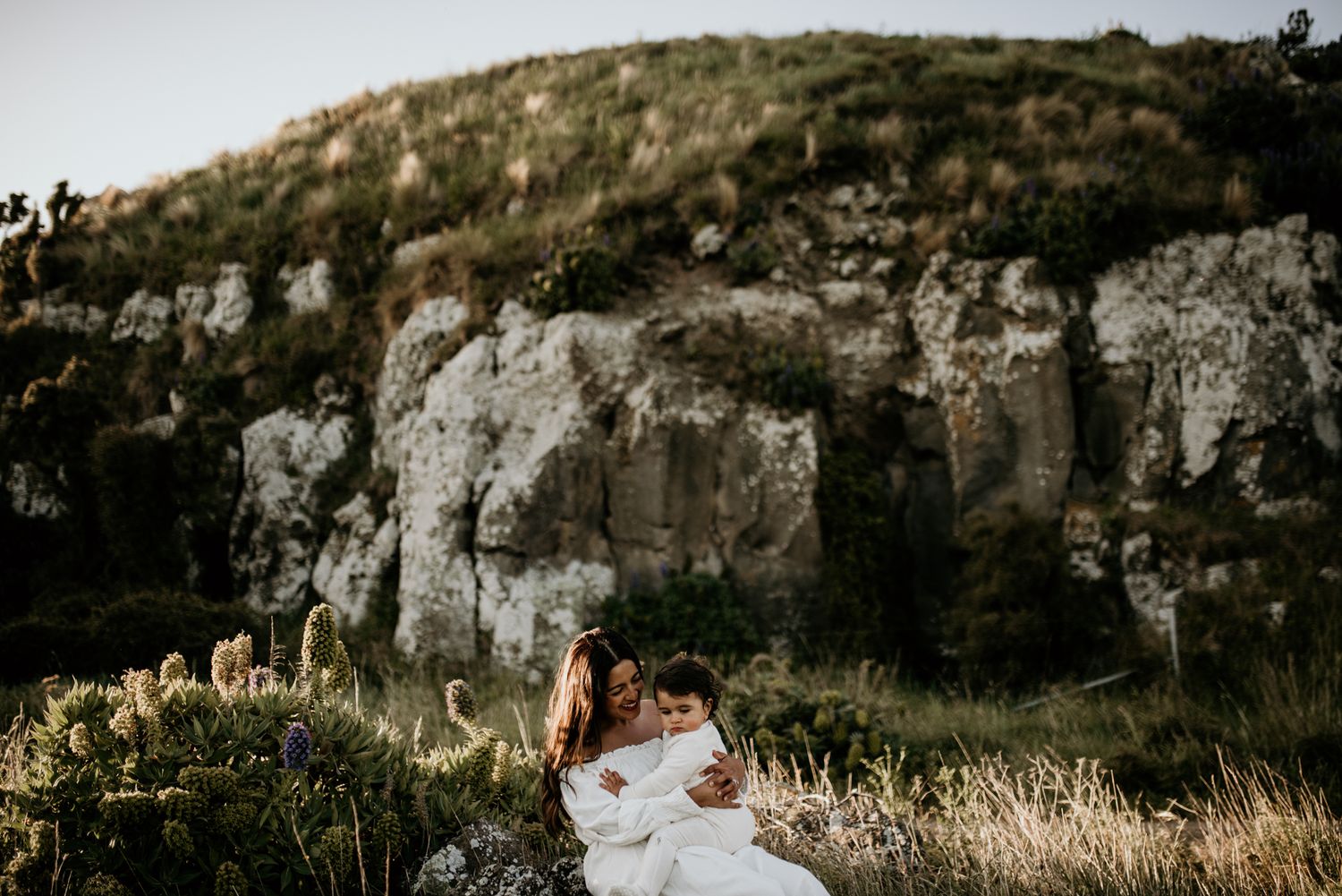 A person in white sits among wildflowers with rocky cliffs in the background.