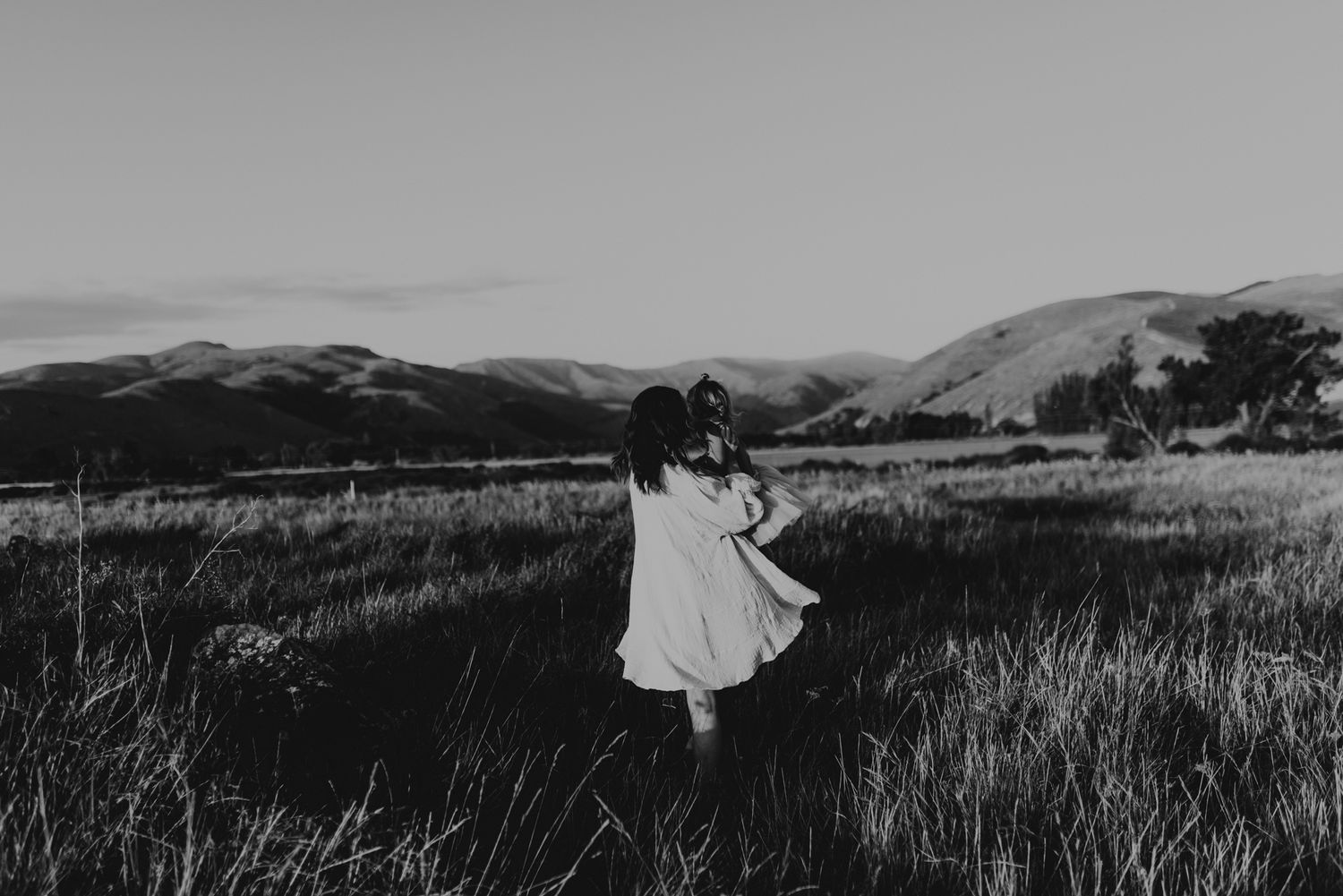Black and white image of two people dancing in a field with mountains in the background.